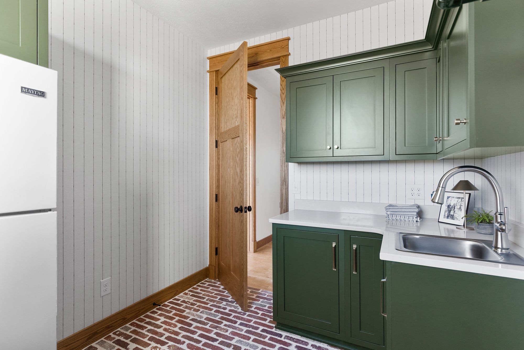 Laundry room featuring green cabinetry, white countertops, and brick flooring. An open wood door reveals another room in the background, with a white Maytag appliance on the left.