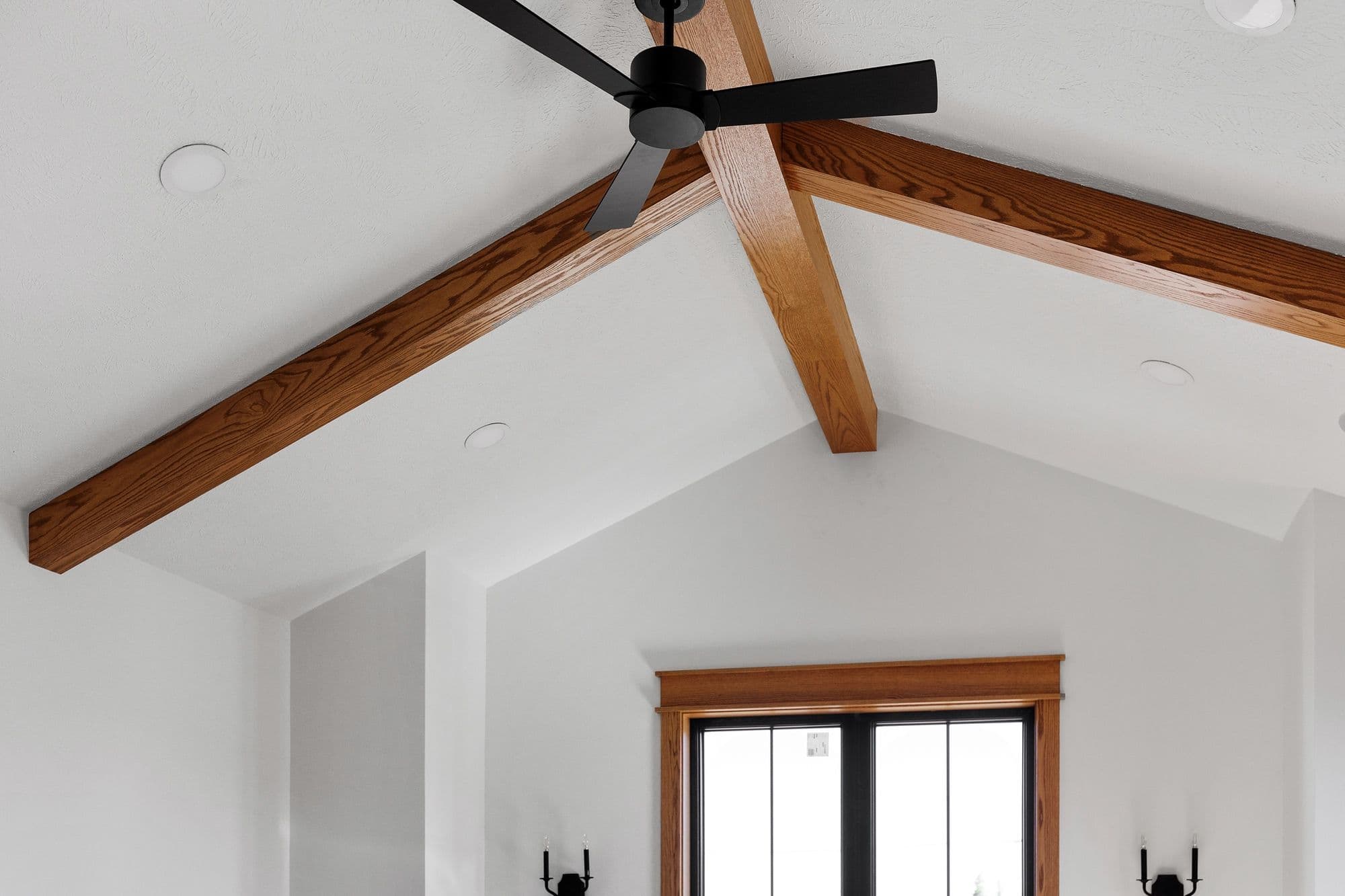 Angled shot showcases a modern living room ceiling with exposed wooden beams and a sleek black ceiling fan. The room features recessed lighting and a window with dark trim.