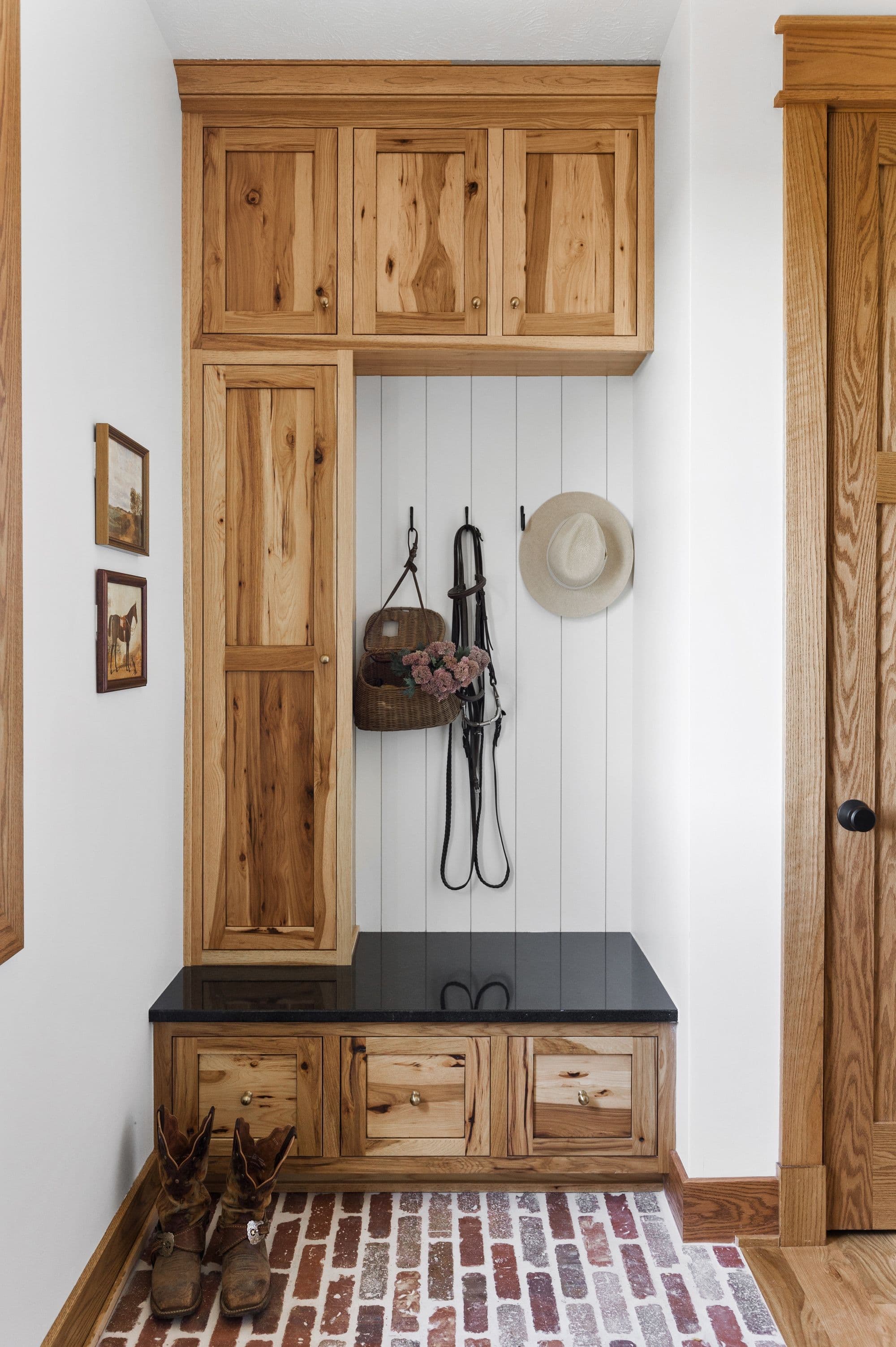Entryway features a built-in mudroom storage system with wooden cabinets and drawers topped with a dark countertop. Cowboy boots sit on a brick-patterned floor, adding rustic charm.