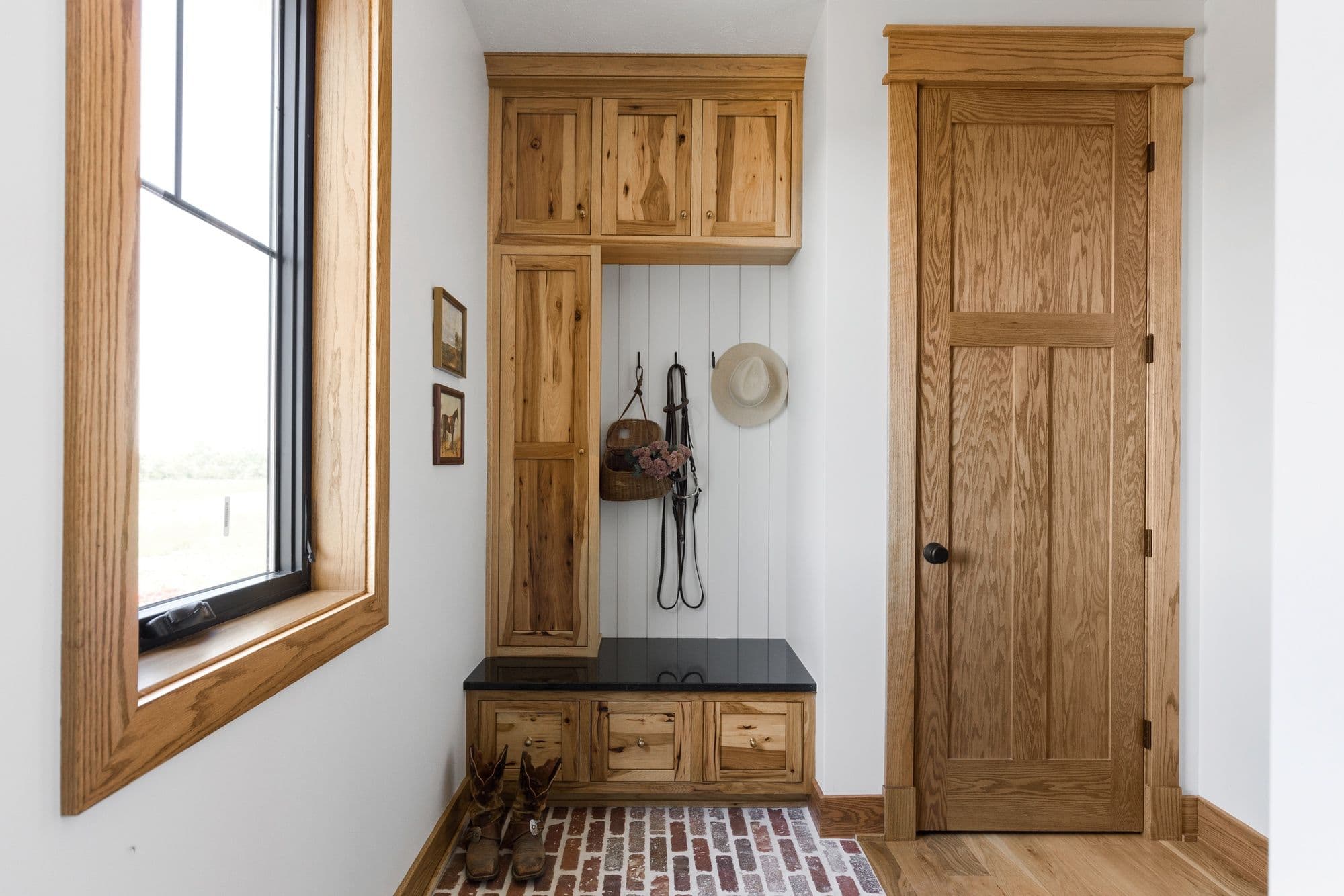 Mudroom features wood cabinetry with upper cabinets, bench seating, and coat hooks. The space includes brick flooring, a window with wood trim, and a wooden door.