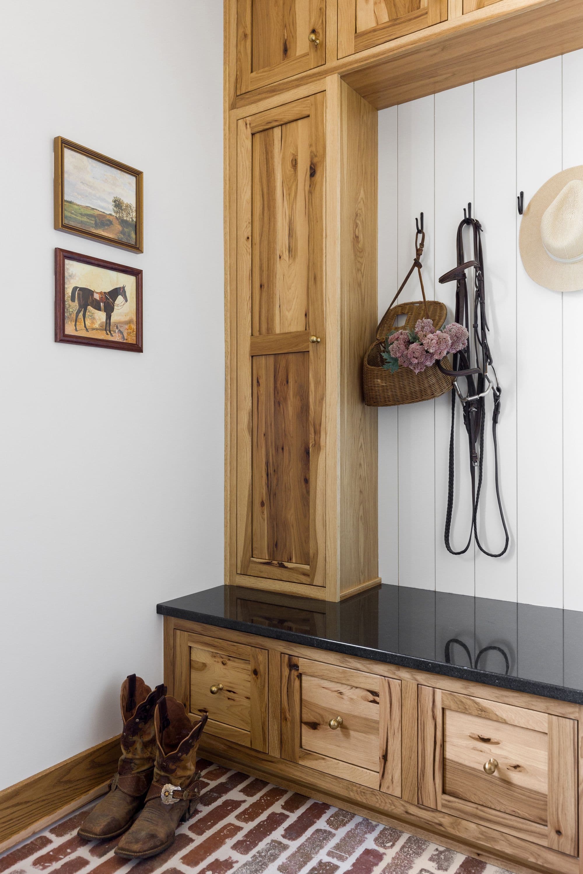 Mudroom with custom wood cabinetry, black countertops, and brick flooring. The space features wall hooks with equestrian accessories and artwork displayed on the wall.