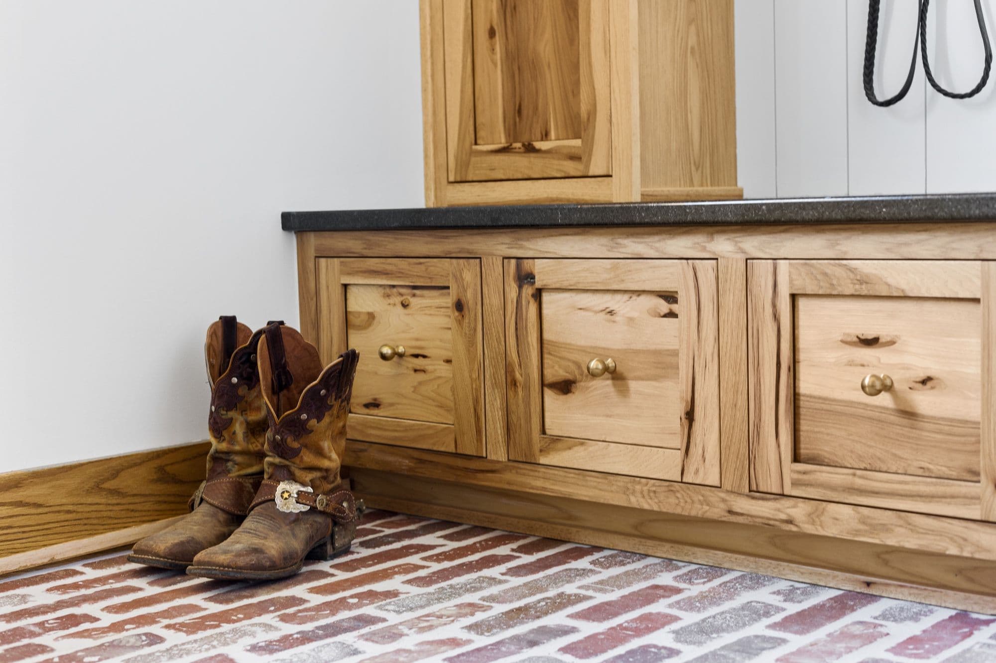 Entryway featuring light wood cabinetry with brass hardware and a dark countertop, paired with cowboy boots on a brick floor. A small matching cabinet sits on top of the countertop.