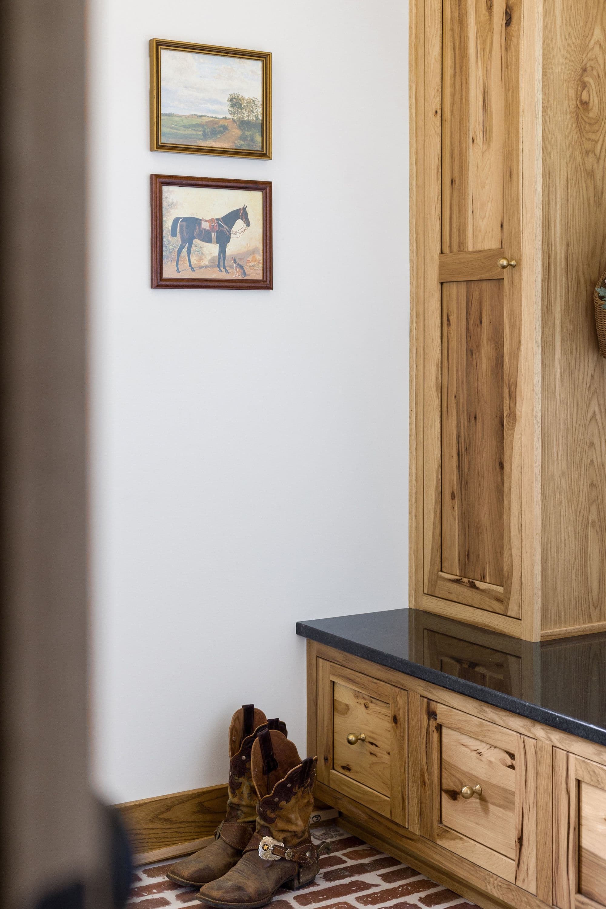 Entryway with wooden built-in cabinets, black countertop, cowboy boots, and framed artwork on the wall. The floor is made of brick.
