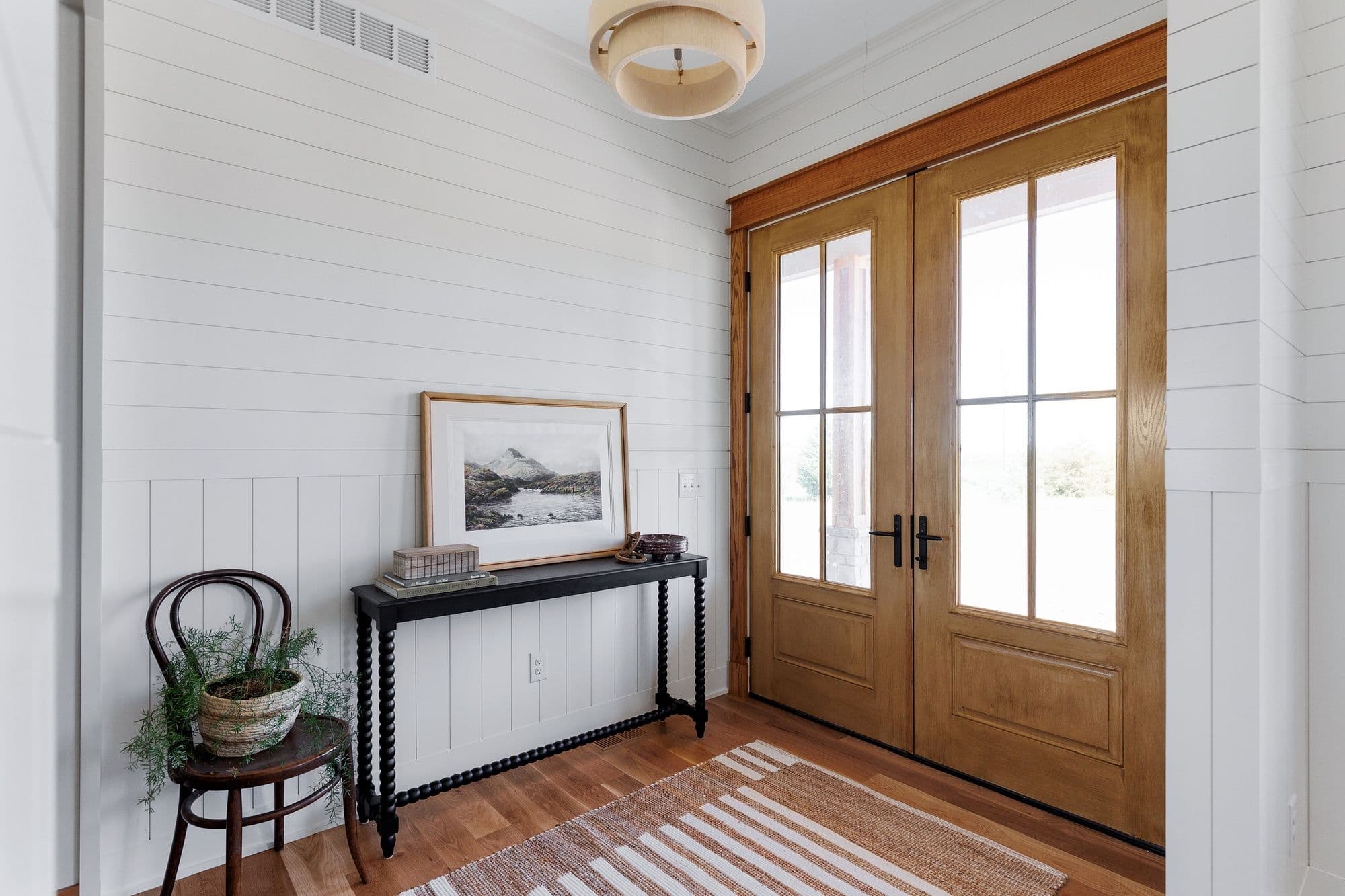 Entryway features light wood double doors with glass panes, opening into a home with white shiplap walls and hardwood floors. A black console table with a framed landscape artwork and decorative items sits against the wall, complemented by a wooden chair with a potted plant.