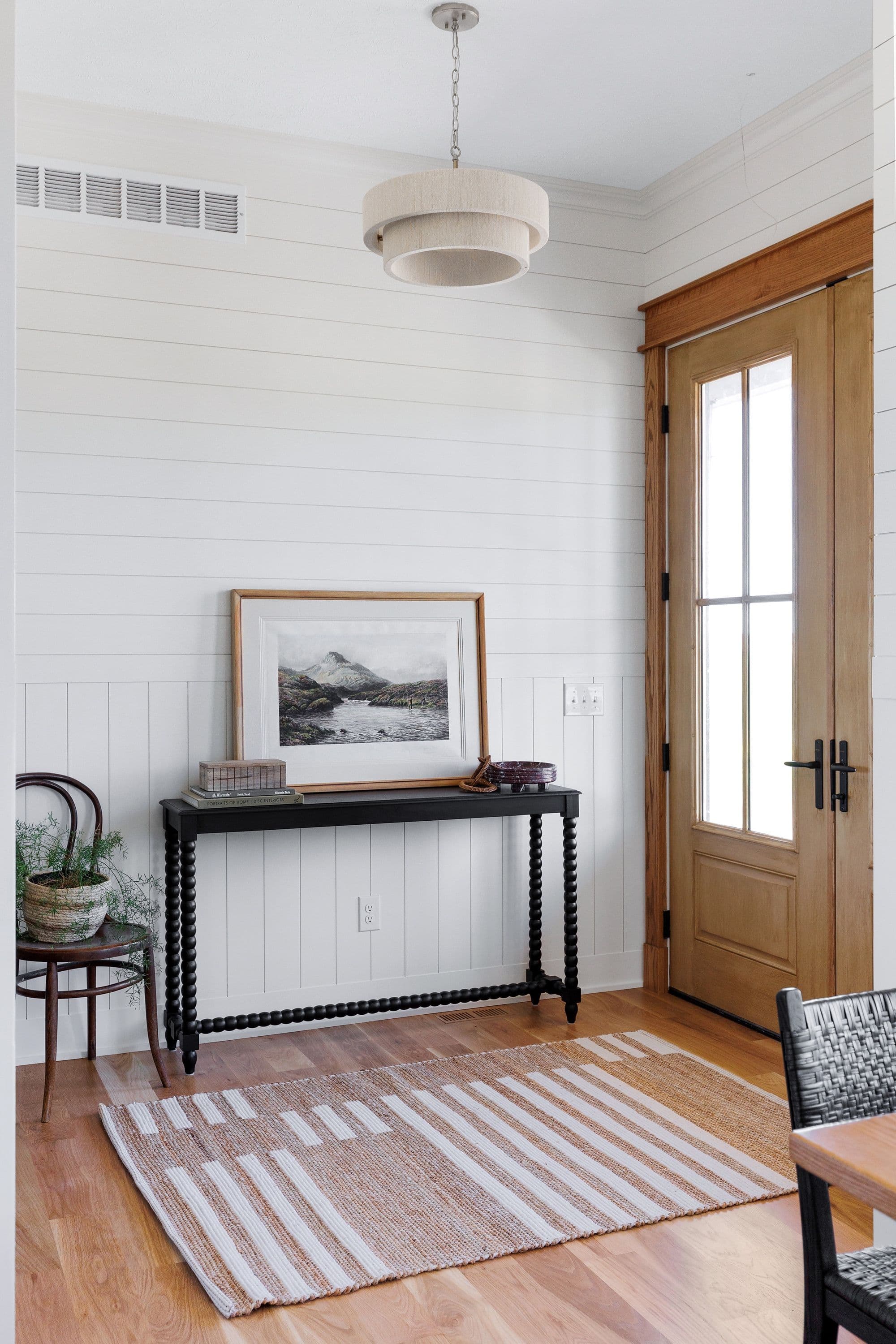 Entryway with white shiplap walls and a wooden door with glass panels. A black console table with a framed landscape print sits against the wall, complemented by a striped area rug.