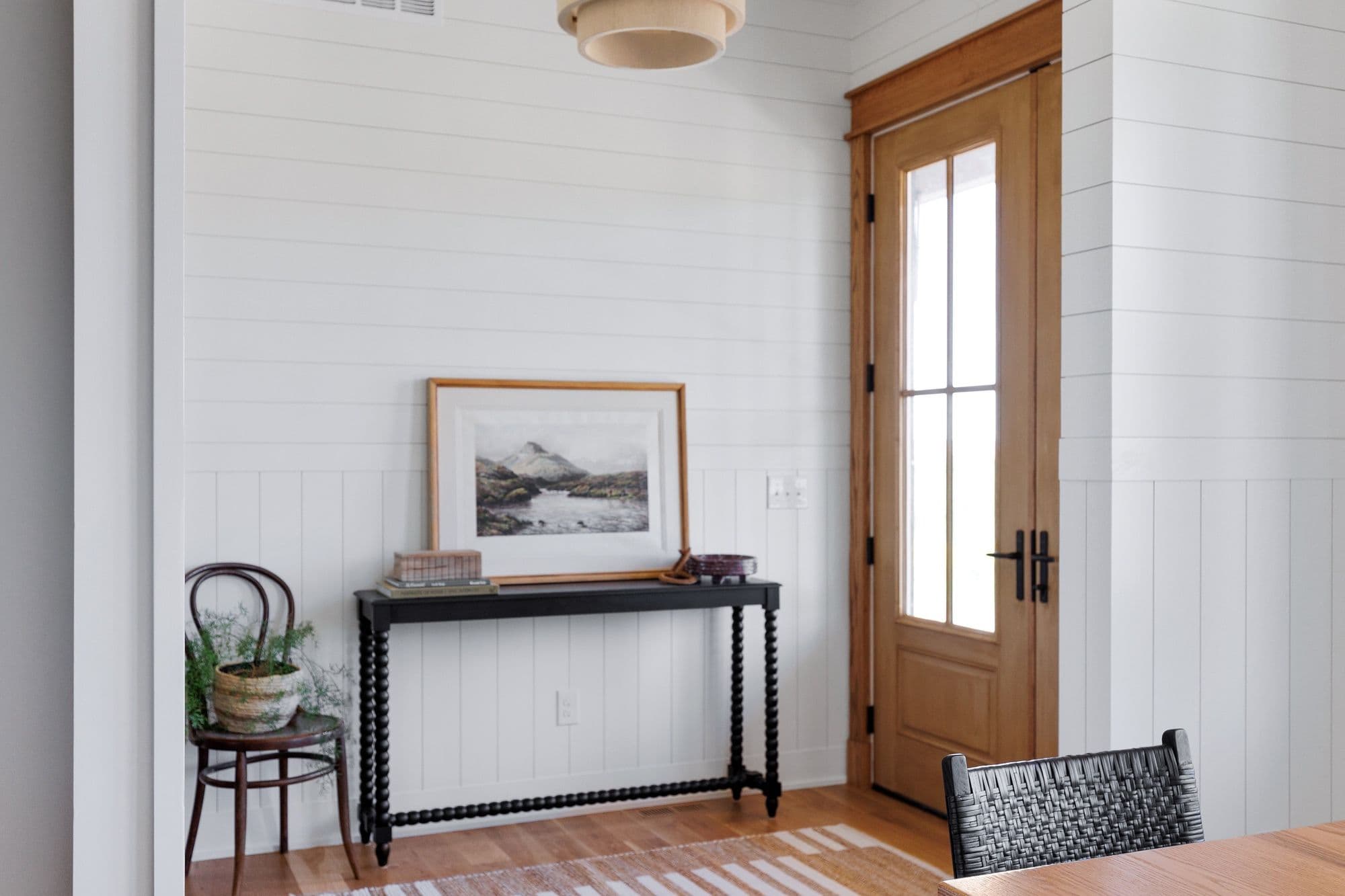 Entryway with white shiplap walls, a black console table with a framed landscape painting, and a wood-framed glass paneled door. A bentwood chair with a potted plant adds a touch of greenery.