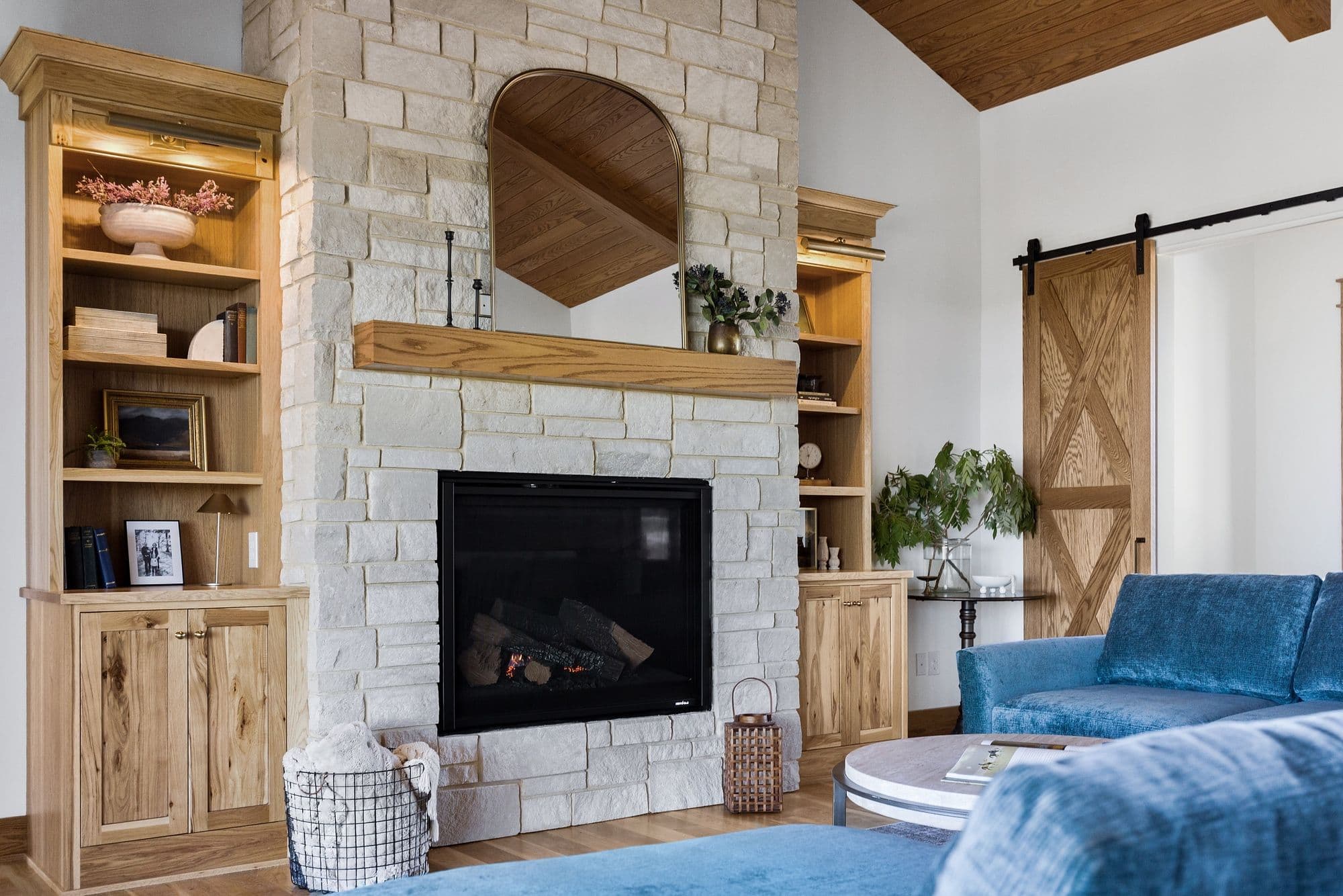 A cozy living room features a stone fireplace with a wooden mantel and arched mirror above. Built-in wooden bookshelves flank the fireplace. The room includes a blue sofa, wooden floors, and a sliding barn door, creating a warm and inviting atmosphere.