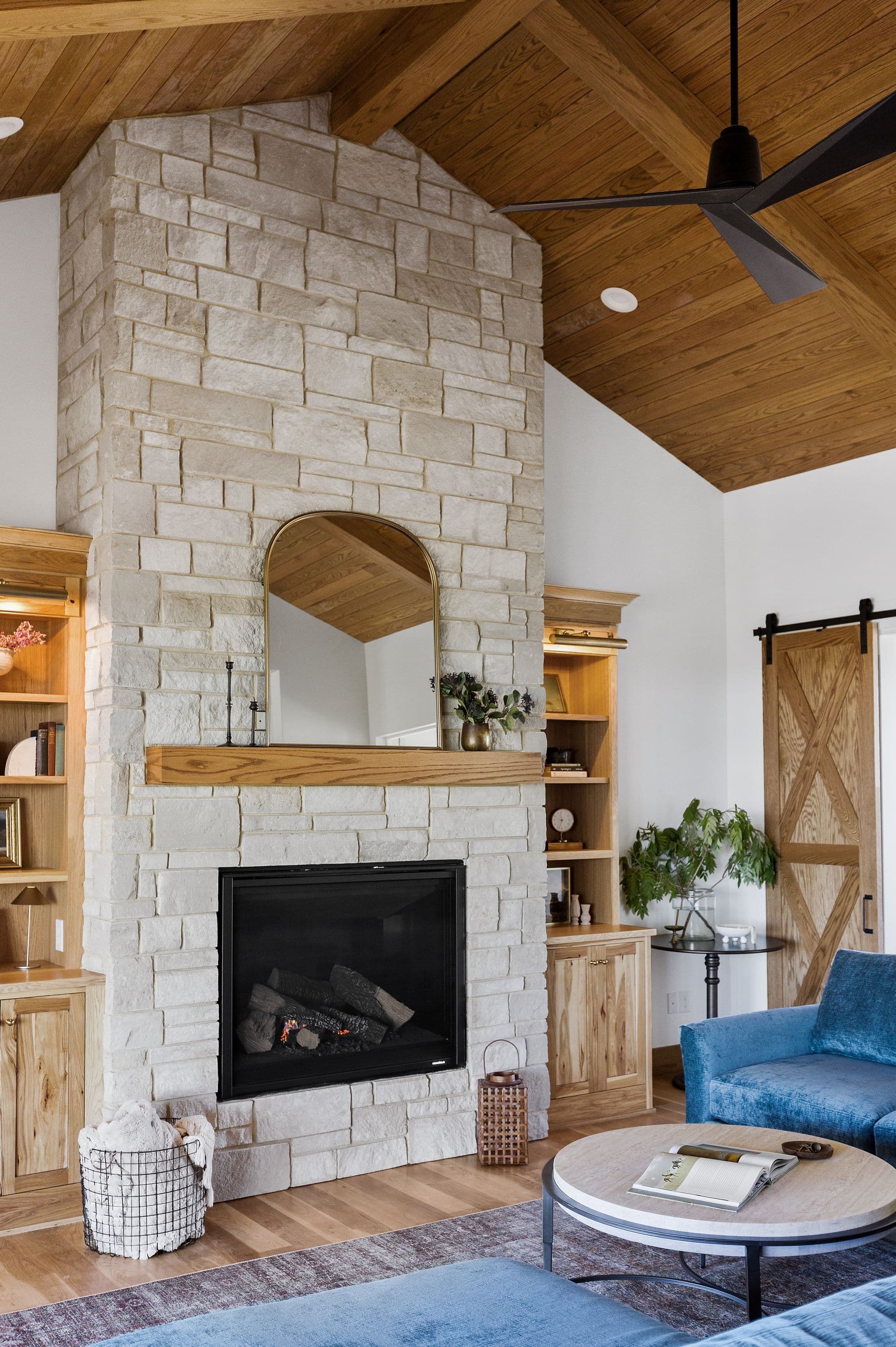 Living room showcasing a stone fireplace with wooden mantel, flanked by built-in shelves. The room has a vaulted ceiling with wood paneling and a black ceiling fan, complementing the barn door and blue velvet furniture.