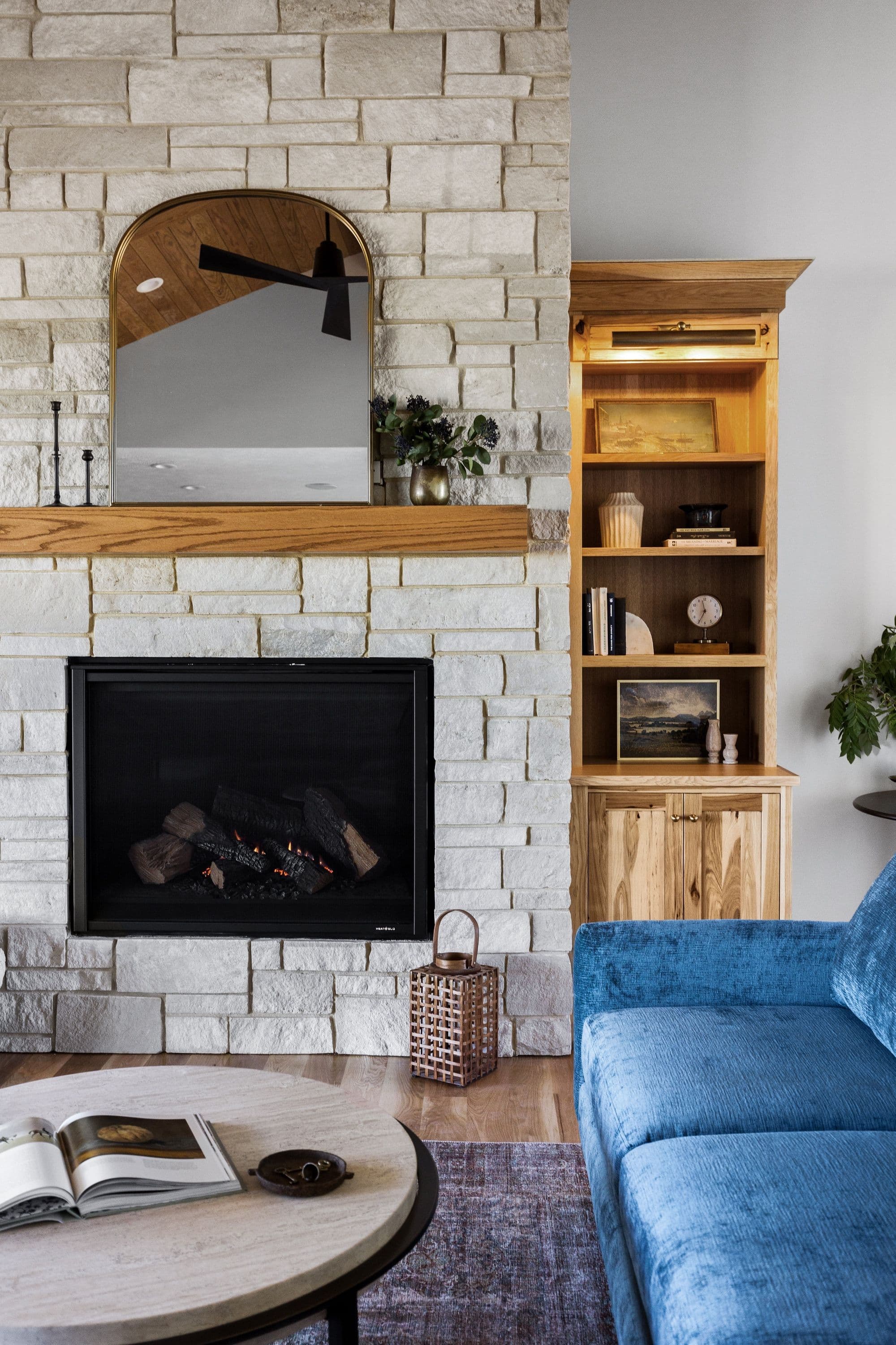 The image shows a cozy living room with a stone fireplace and built-in bookcase. The fireplace has a wooden mantel and a mirror hanging above it, with a blue sofa and round coffee table in the foreground.