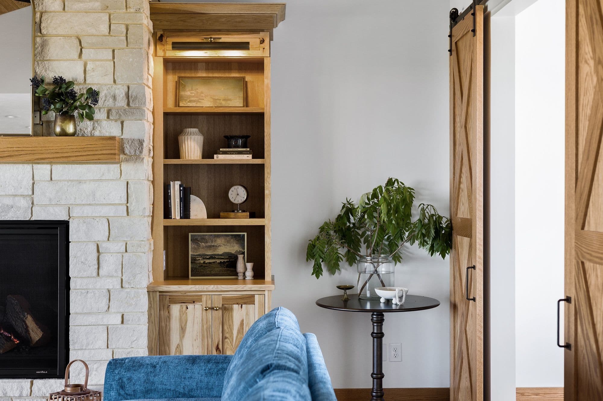 Living room interior with stone fireplace, built-in shelving, and a wooden barn door. The room features a round side table with a plant, and a blue upholstered sofa in the foreground.