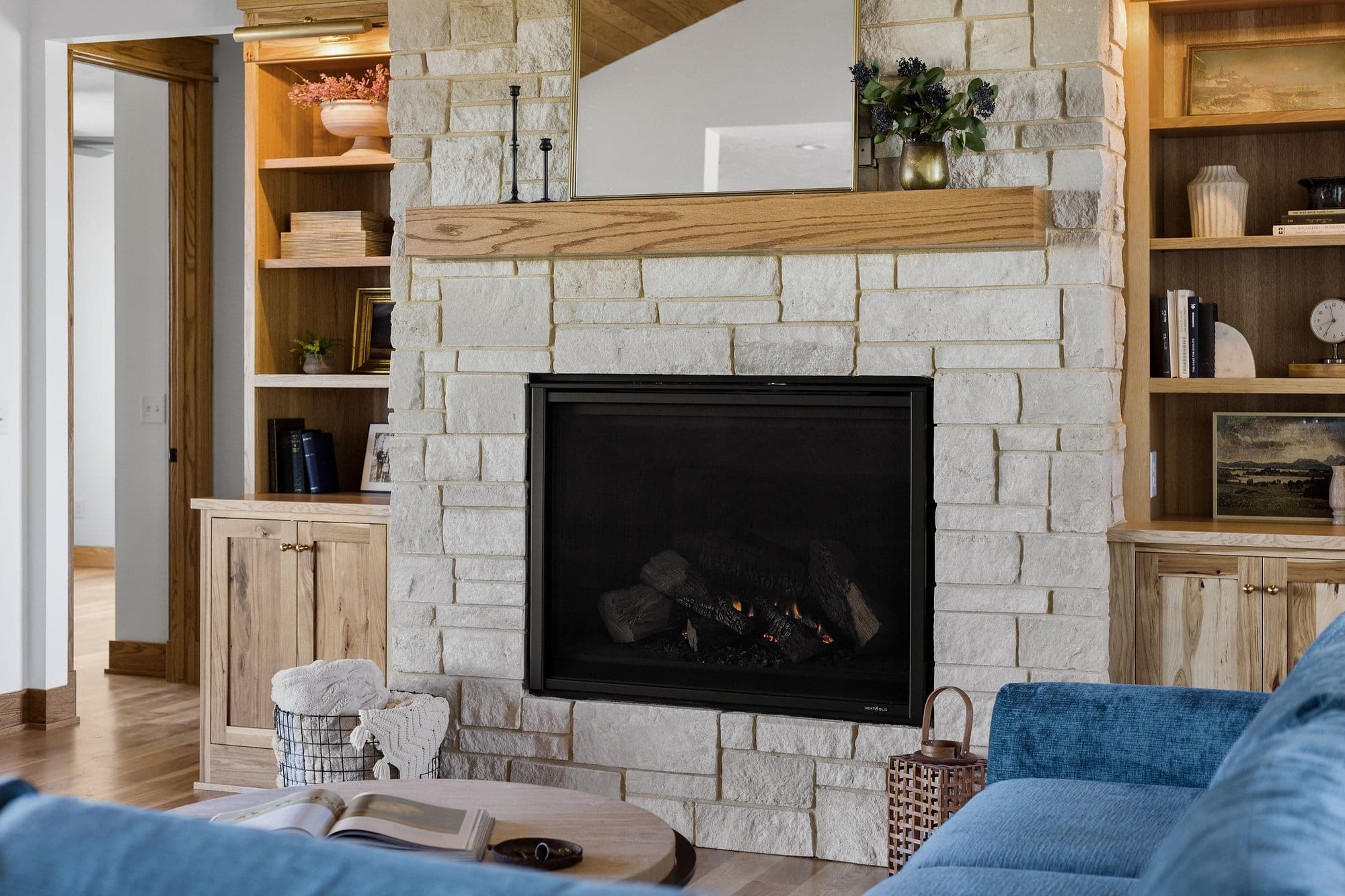 Living room featuring a stone fireplace with a wooden mantel and a black firebox, flanked by built-in wooden cabinets and bookshelves. The space is illuminated by warm lighting, and a blue couch is positioned in front of a round wooden coffee table.