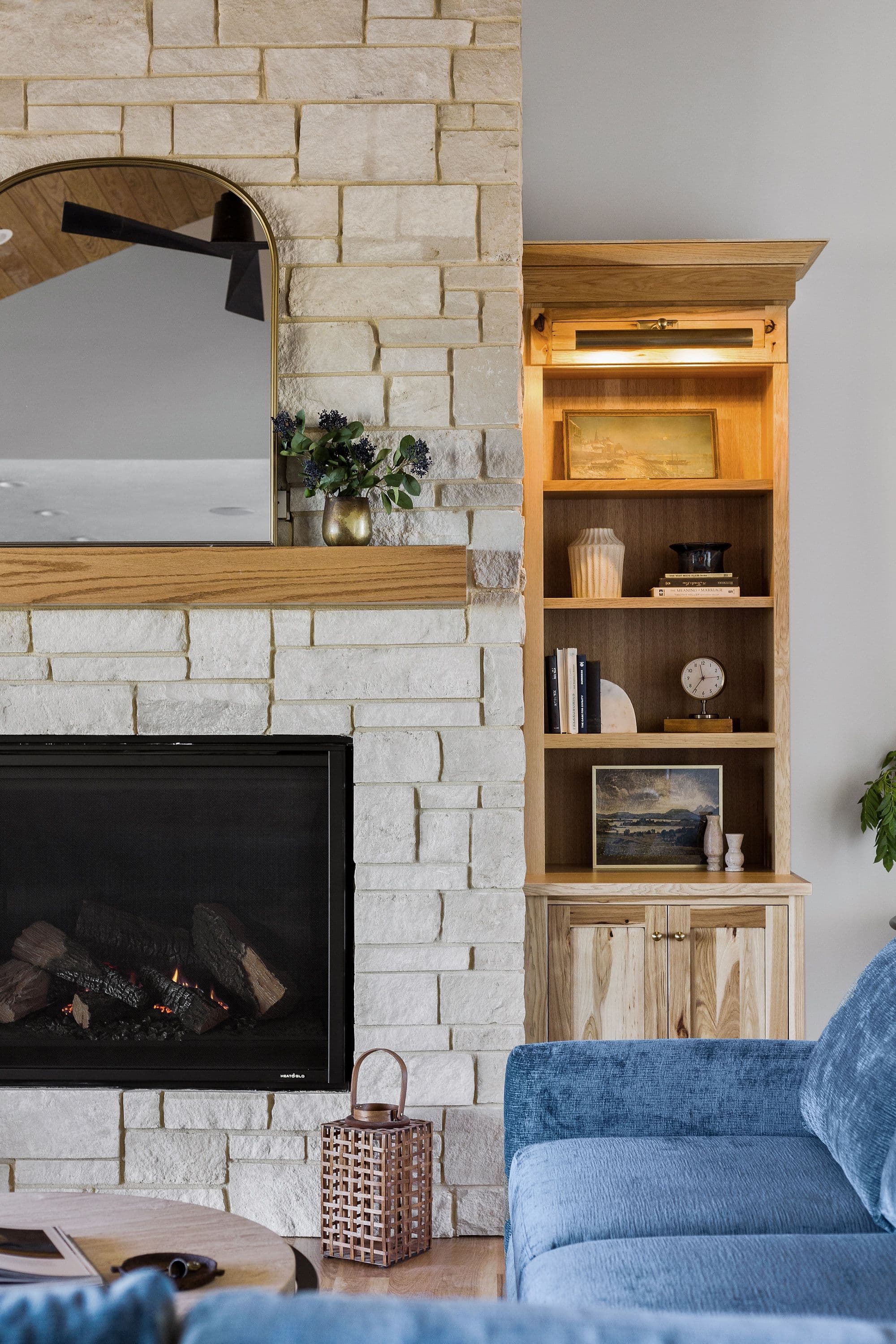 This living room features a stone fireplace with a wooden mantle and a gold-framed mirror, complemented by a light wood built-in bookshelf. A blue velvet sofa sits adjacent to the bookshelf, creating a cozy and stylish interior.