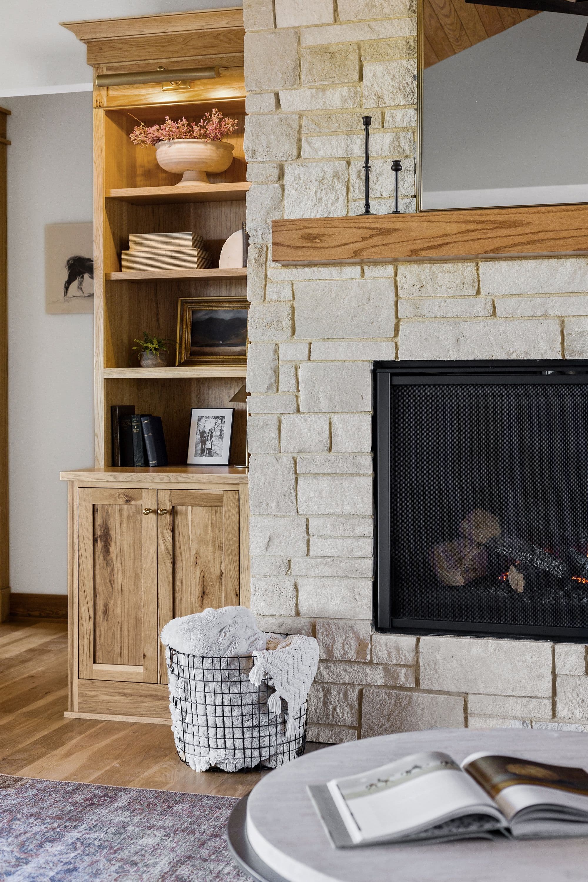 Living room features a stone fireplace with a wooden mantel, built-in wooden shelving with display items, and a wire basket filled with a soft blanket. The room also has hardwood flooring and a round coffee table with a book.