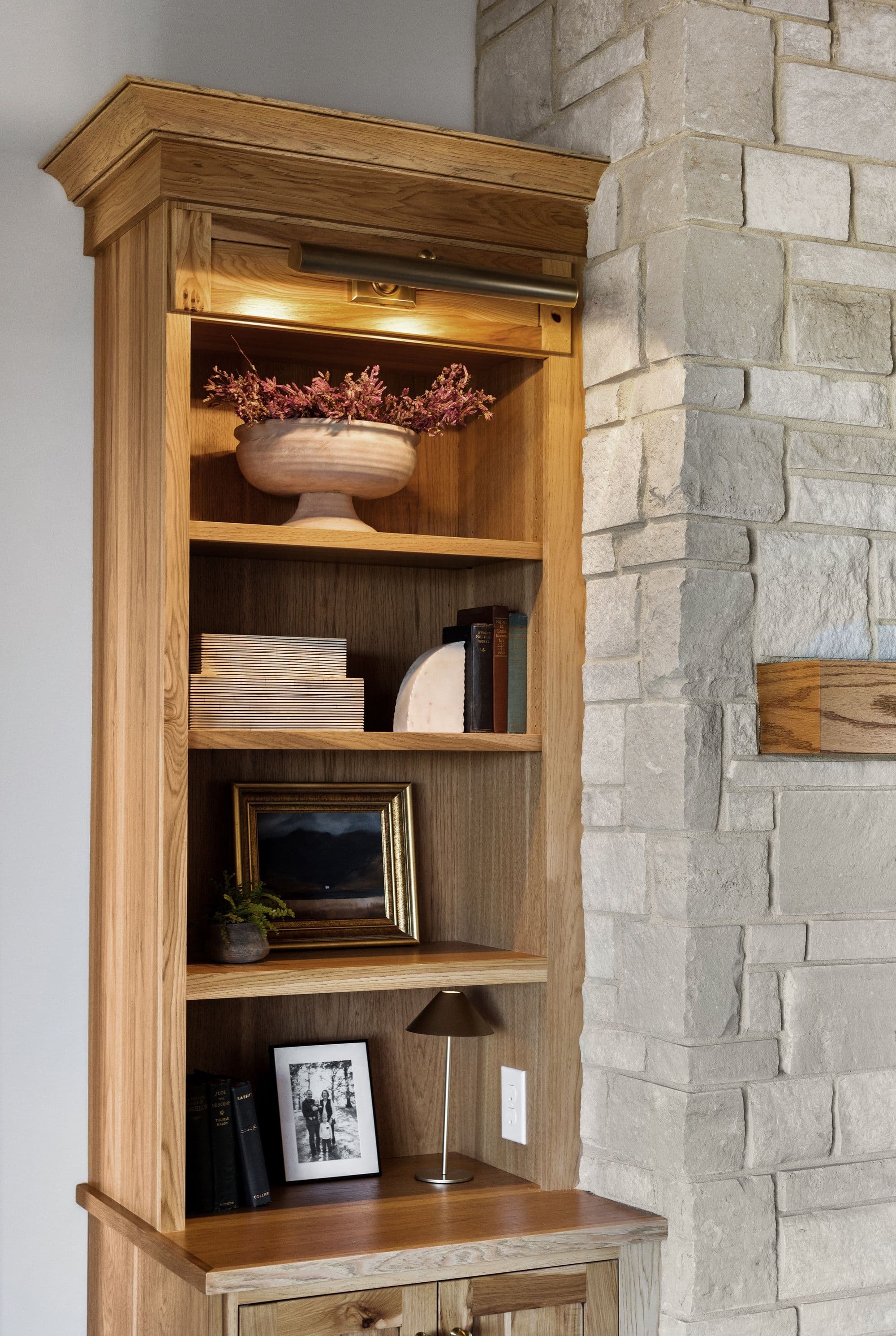 Built-in wooden bookshelf with a lamp and a variety of decor, including books, a picture frame, and a vase of flowers. The bookshelf is adjacent to a stone wall.