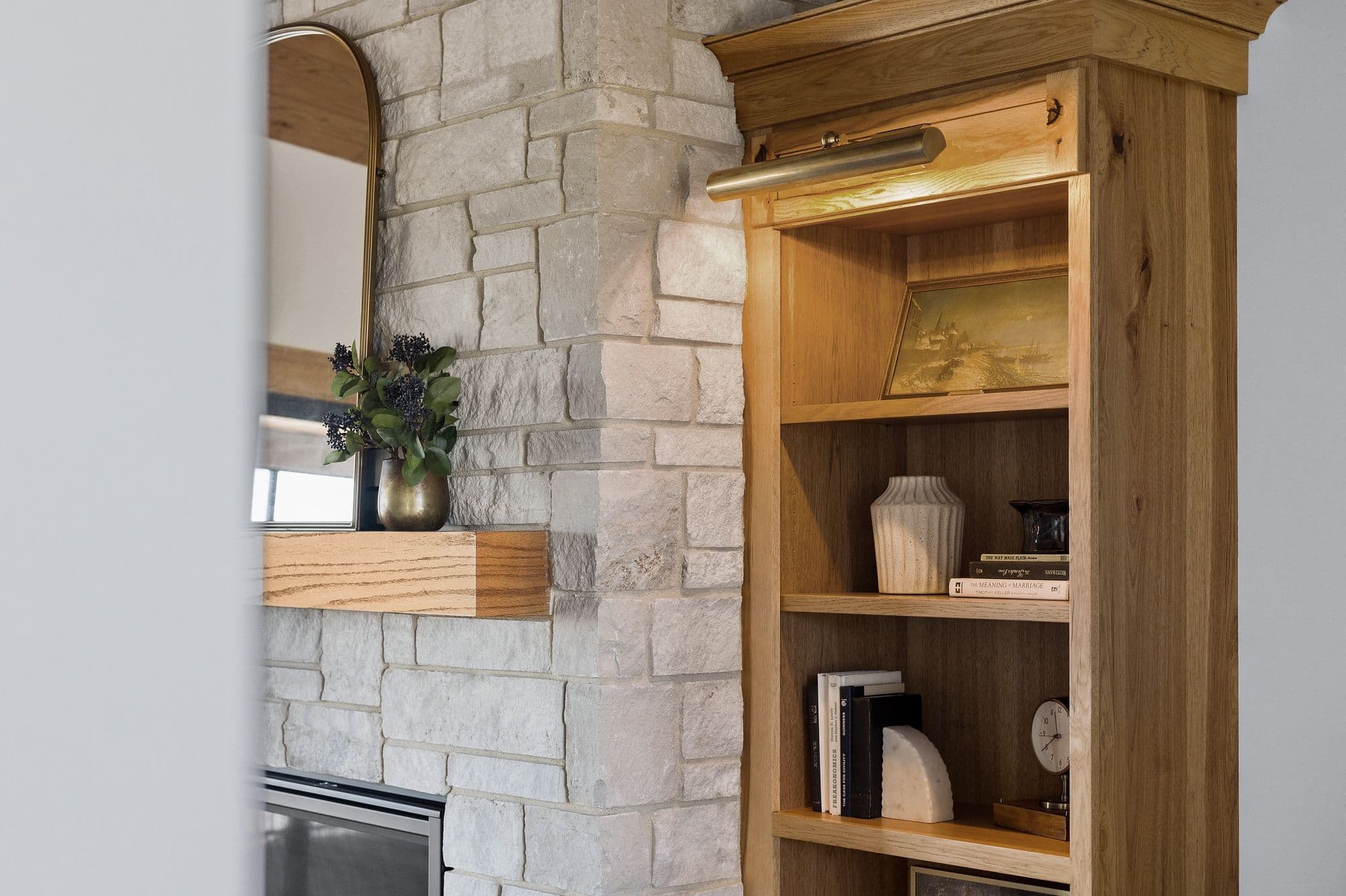 Close-up of a living room featuring a stone fireplace with a wooden mantel, an arched gold mirror, and a wooden bookcase filled with books and decorative items. The room has a cozy and inviting atmosphere.