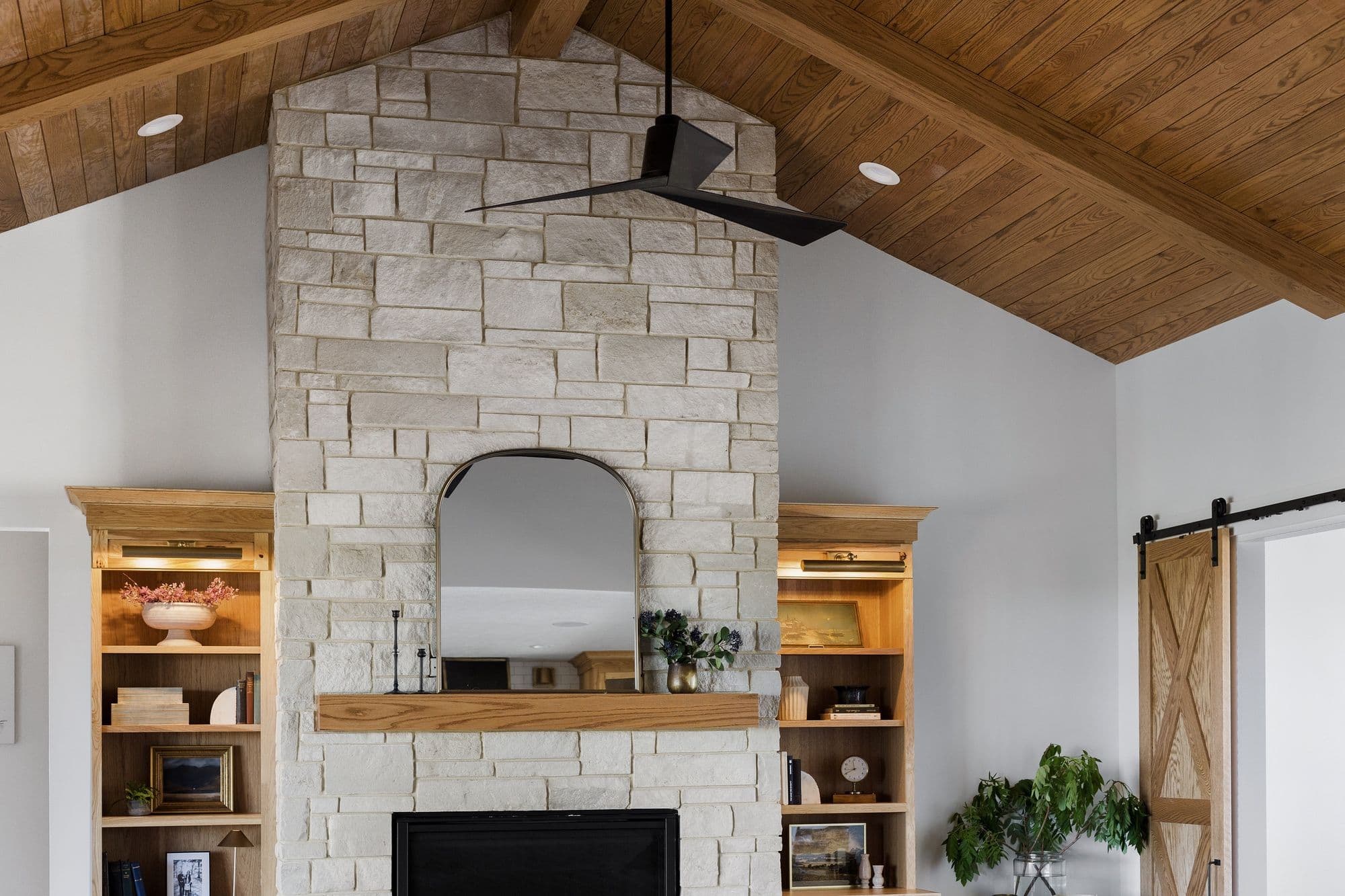 Living room features a stone fireplace with a wooden mantel and a black arched mirror, flanked by built-in wooden bookshelves. The room also has a wood-paneled vaulted ceiling with a black ceiling fan and recessed lighting.