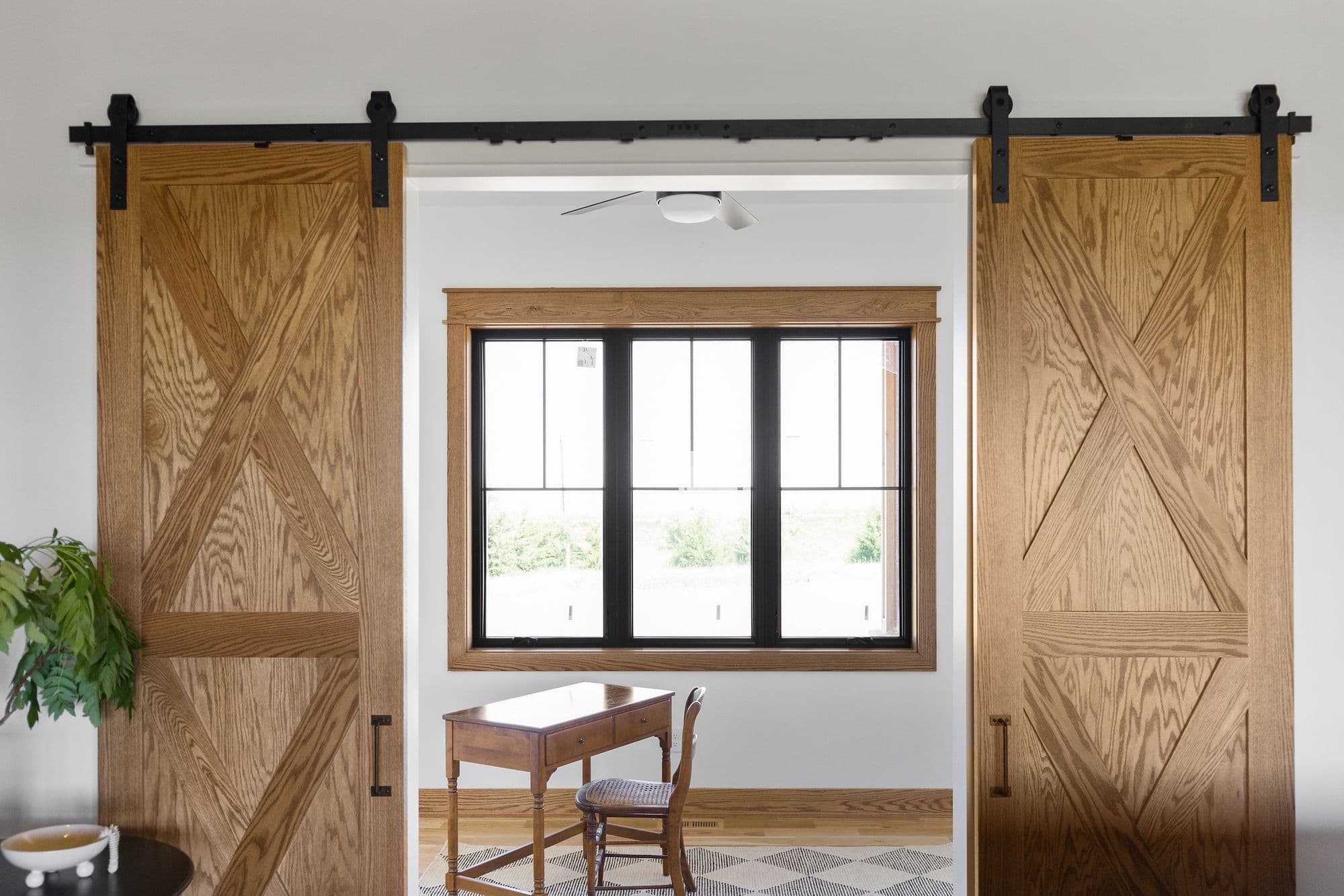 Home office area featuring barn doors, a wooden desk, and a window with a black frame. The room has neutral walls and a patterned rug.