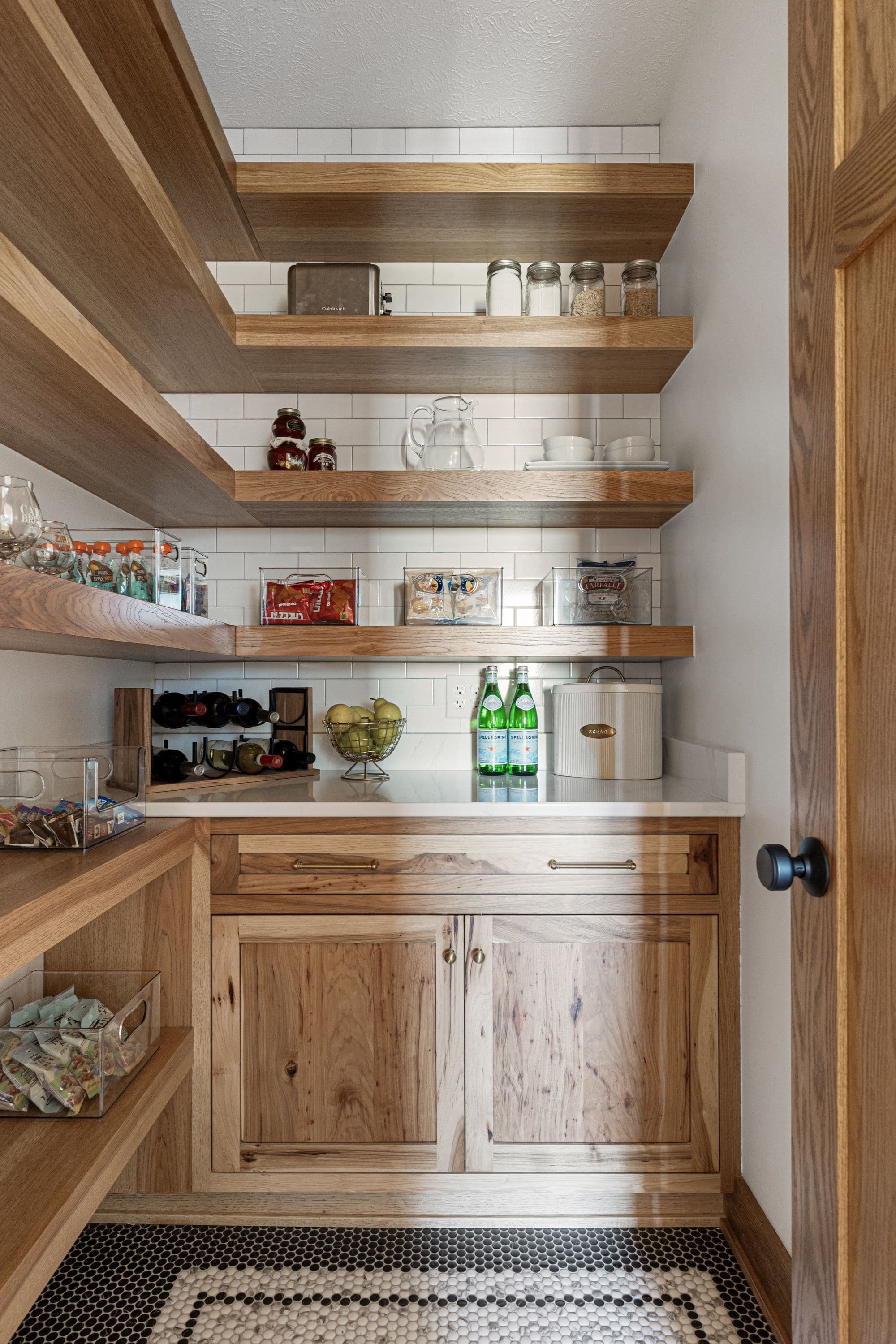 Pantry with light wood shelving and cabinets, white subway tile backsplash, and black and white mosaic floor. The pantry is stocked with a variety of food items and drinks.