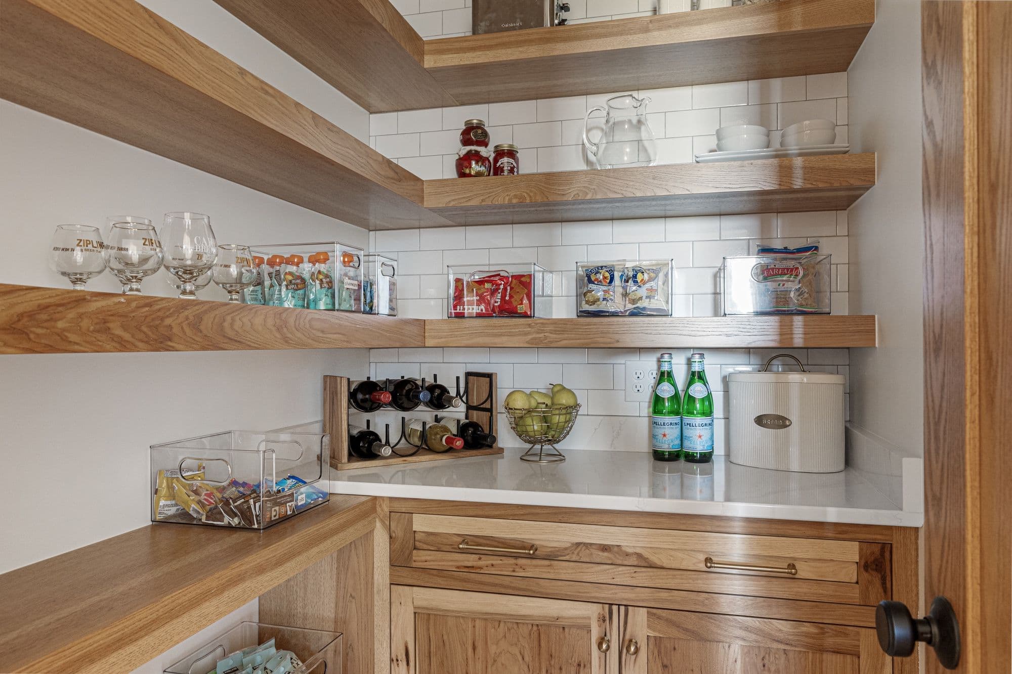 This pantry features wooden floating shelves stocked with glasses, snacks, and pasta. A countertop below holds a wine rack, fruit bowl, and a bread box with wooden cabinetry below.