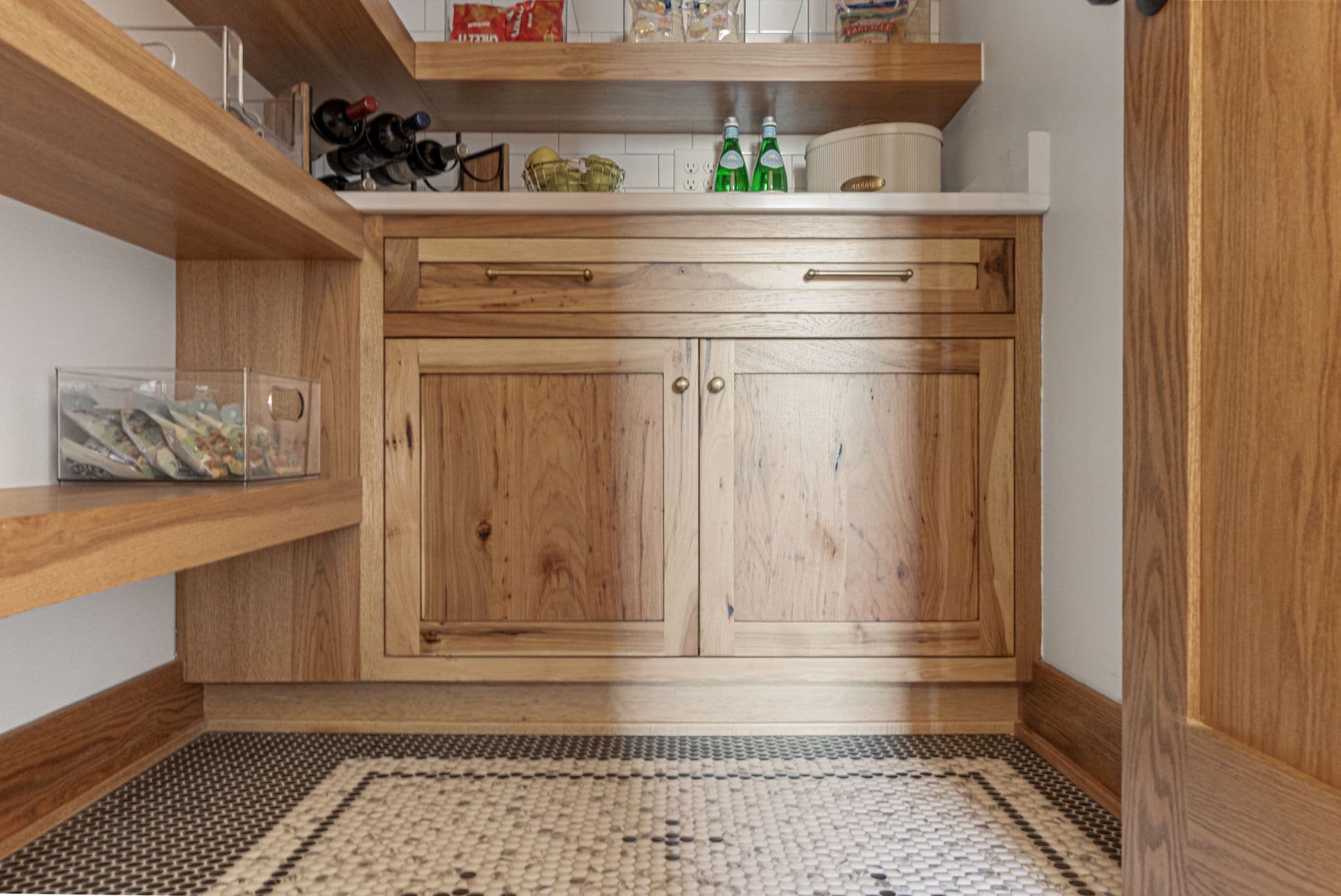 Pantry featuring custom wood cabinetry with inset doors, gold hardware, and open wood shelving. A tile floor with a mosaic pattern adds detail to the room.