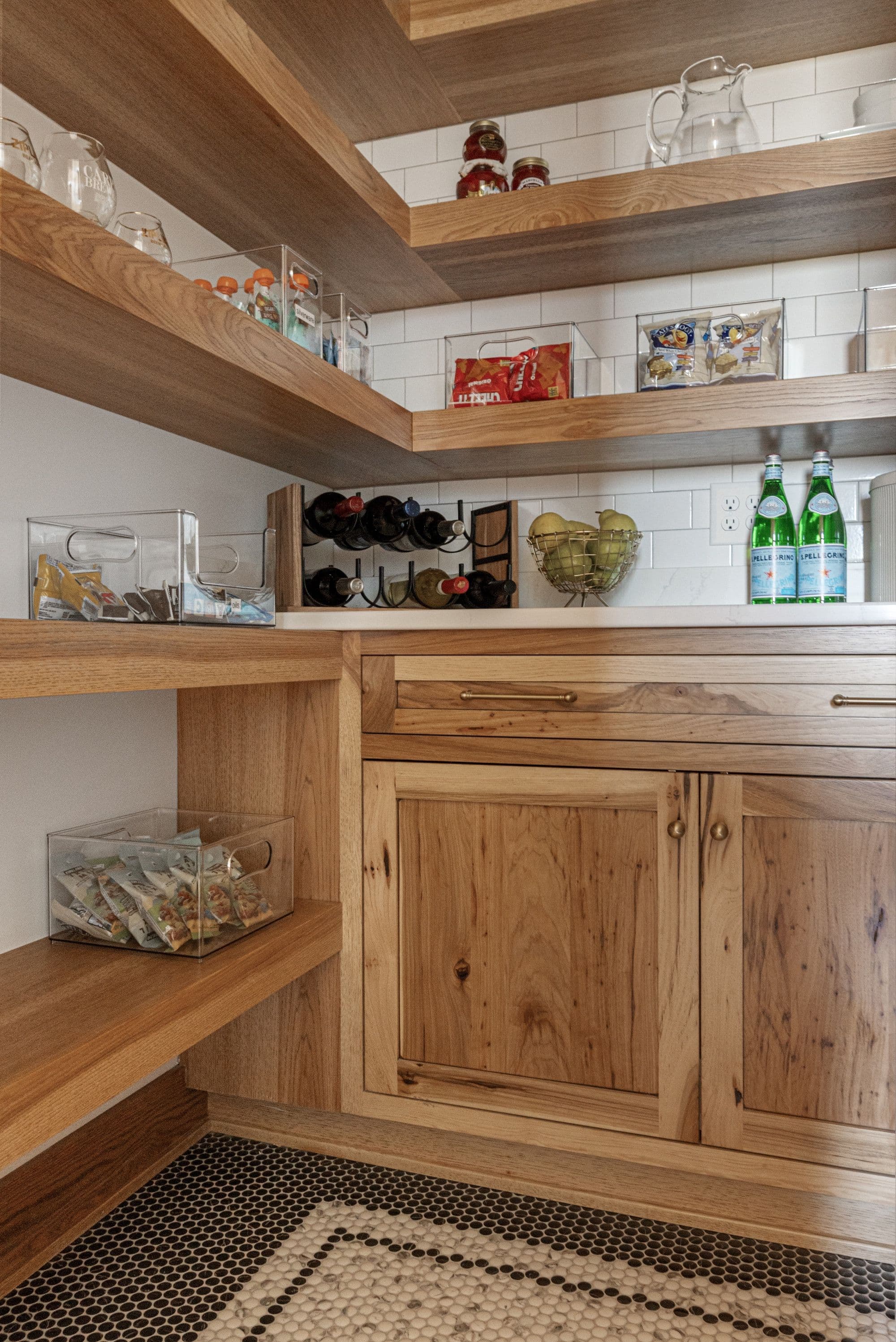 Pantry features wooden floating shelves stocked with snacks and jars, a wine rack, and a metal fruit bowl. The wooden lower cabinetry is accented with gold hardware, and the floor is tiled with small black and white tiles.