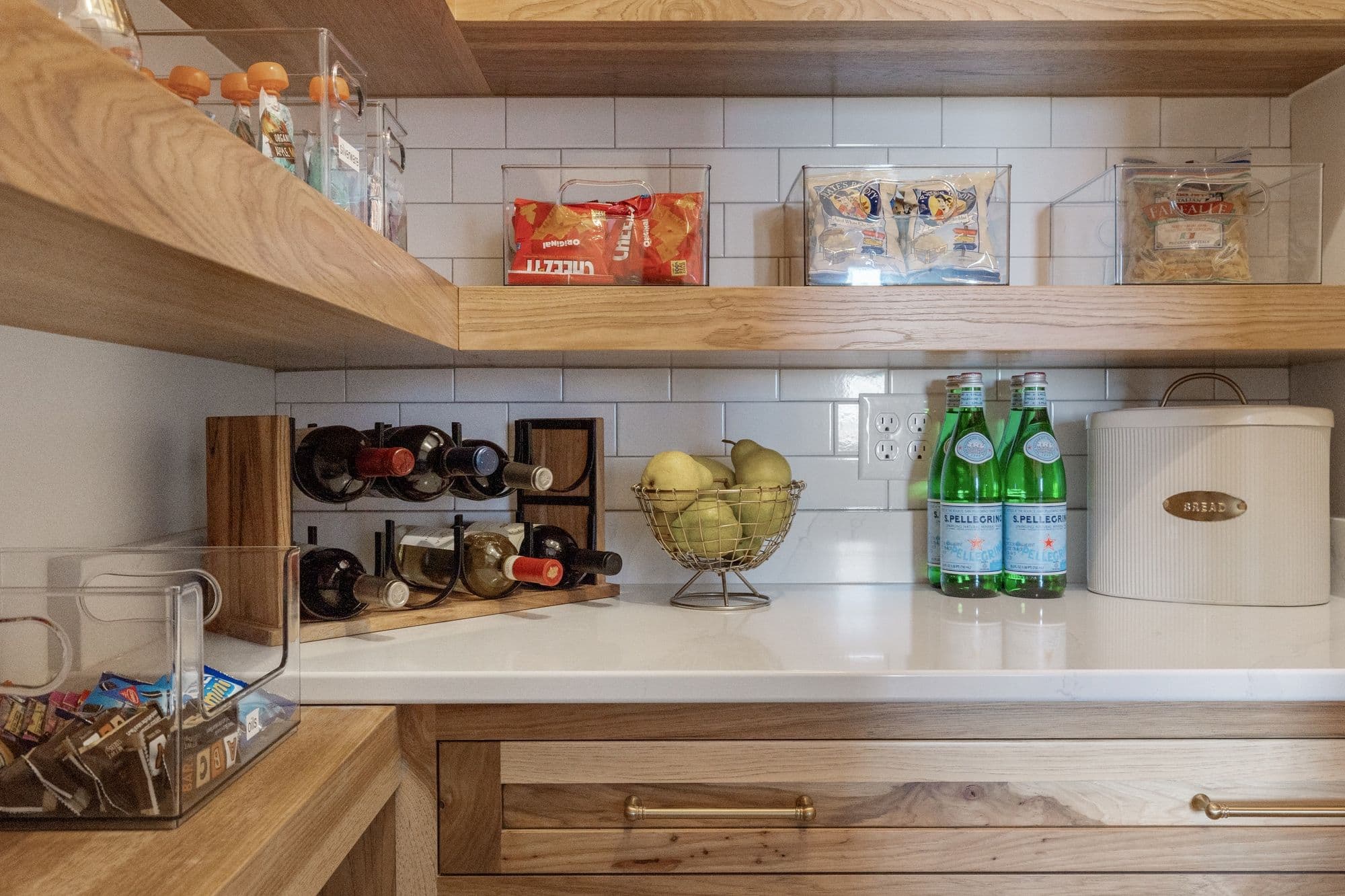 A well-organized pantry is shown featuring light wood open shelving with snacks and water bottles. Clear bins hold pantry staples and snacks while a wire basket holds pears and a wooden wine rack is also displayed.