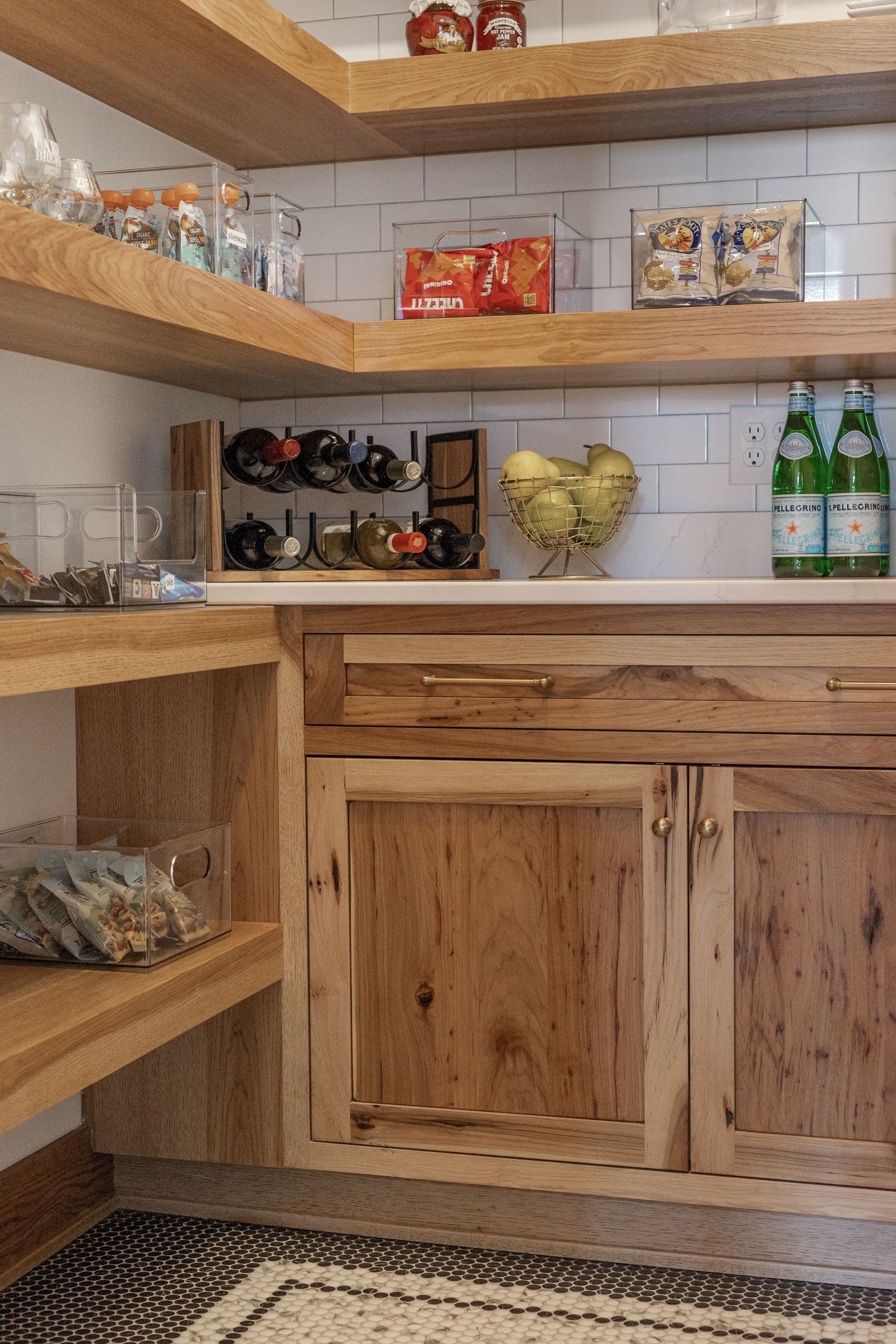 Image showcases a pantry with light wood floating shelves and cabinetry. Clear containers hold snacks, with various jars and wine bottles arranged on the countertop.