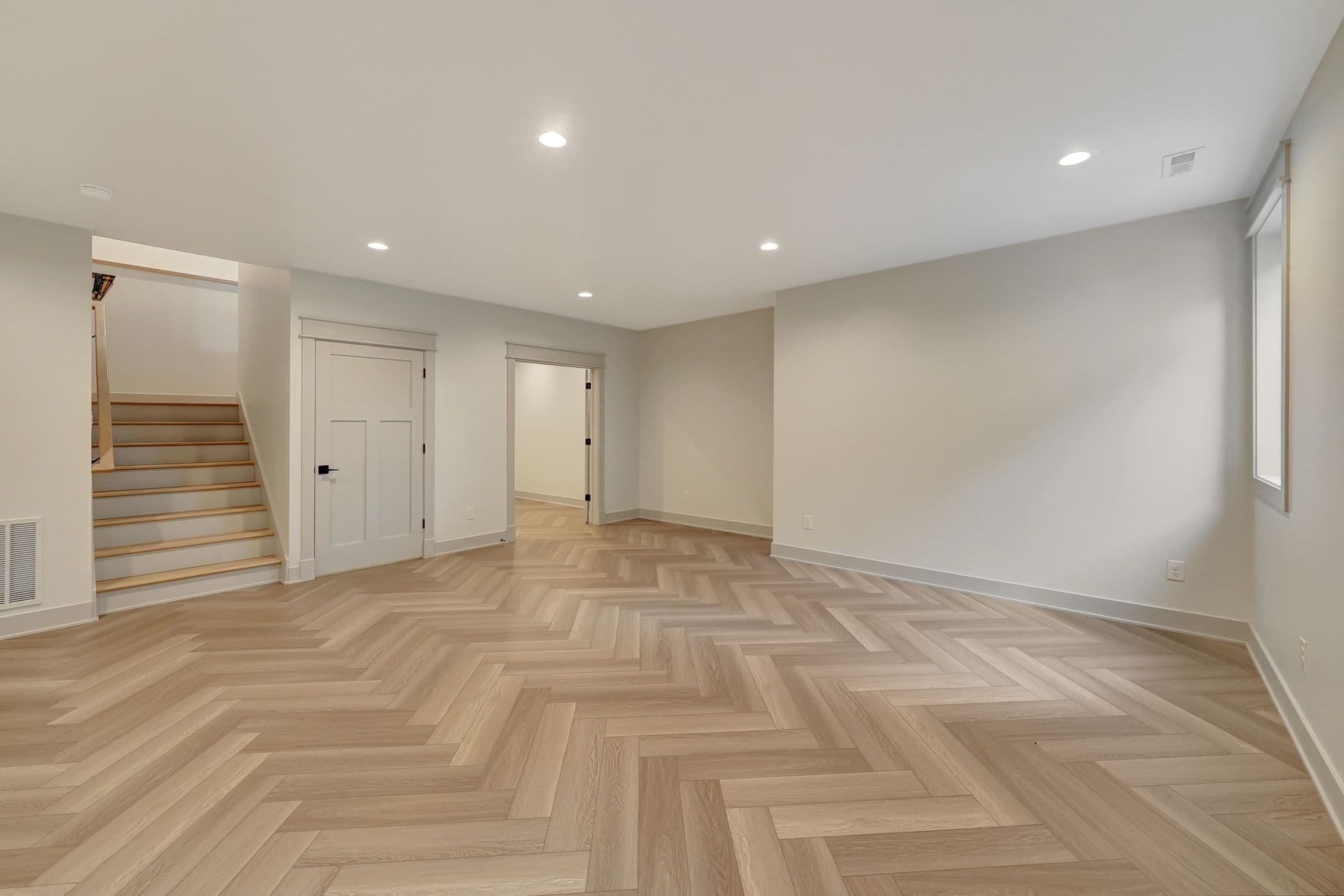 Finished basement space features light herringbone flooring, a staircase leading to another level, and two doorways. The walls are a light neutral color, and recessed lighting illuminates the room.