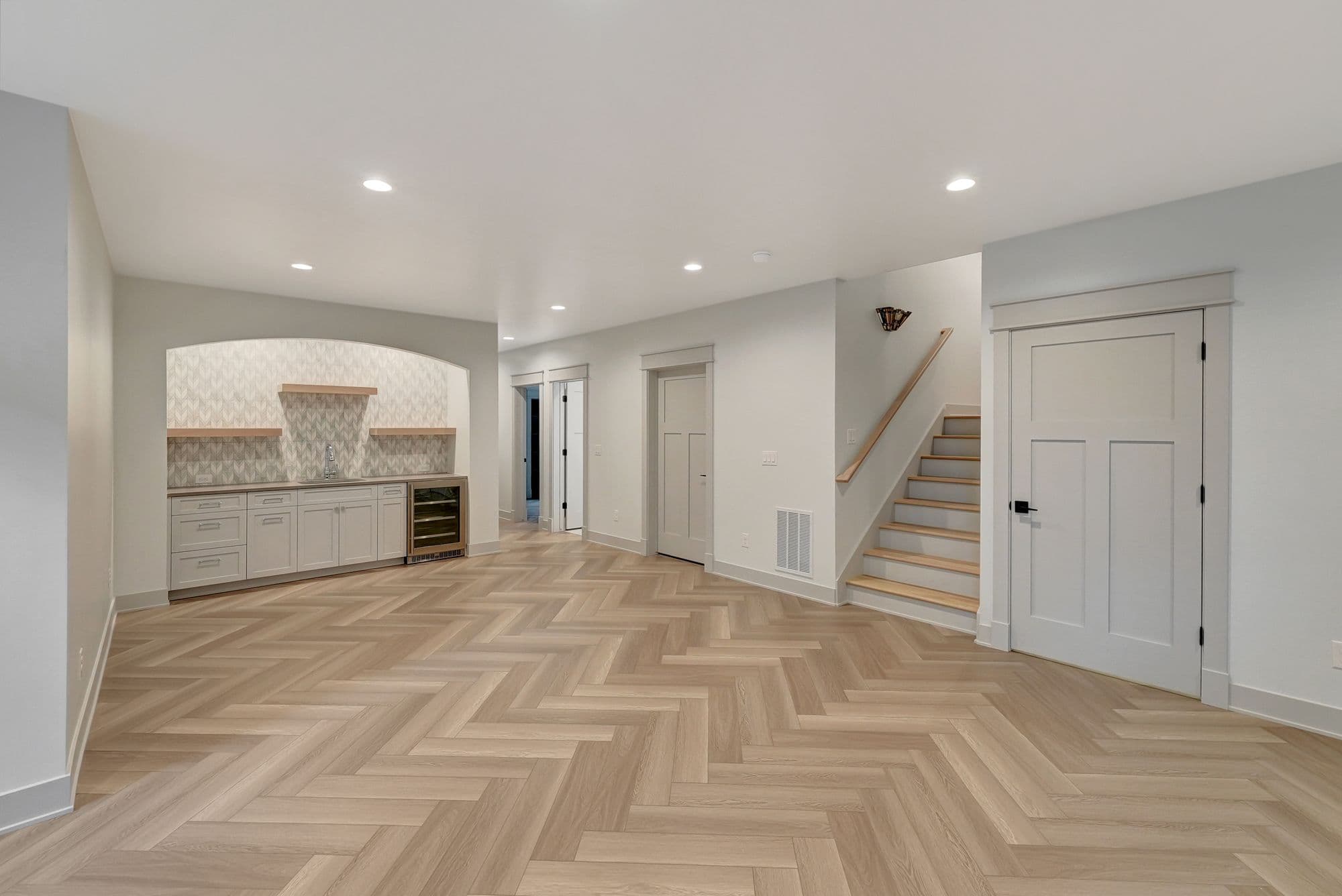 Finished basement with light wood herringbone flooring, built-in bar area with gray cabinets and wine cooler, and white trim. The space also features a staircase with light wood treads, and paneled doors.