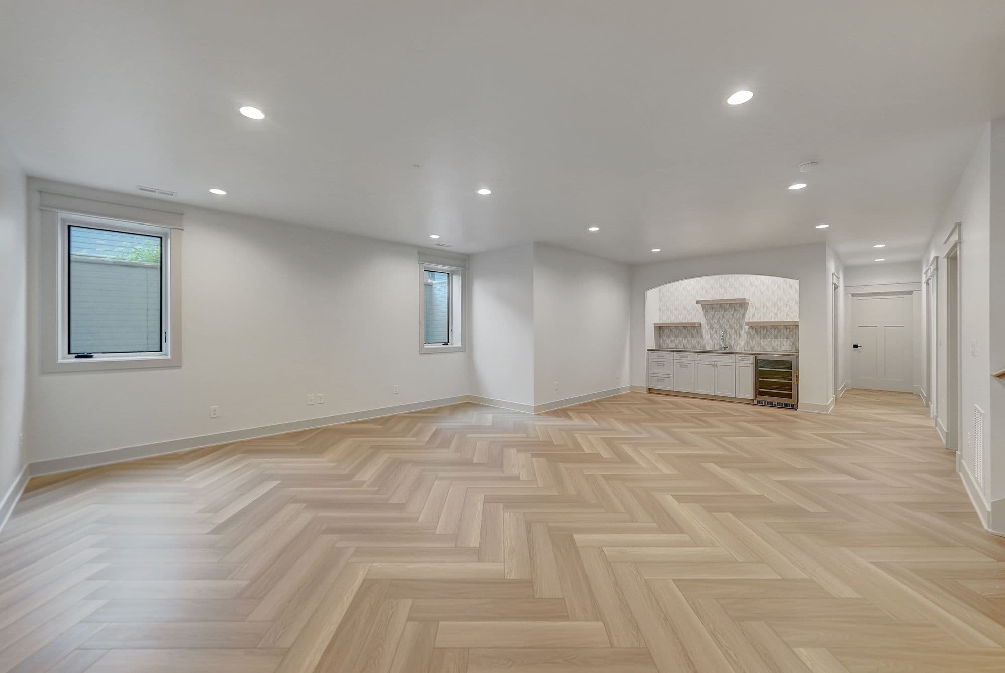 Bright basement features herringbone wood floors, white walls, and recessed lighting. A small bar area includes white cabinets and a wine fridge.