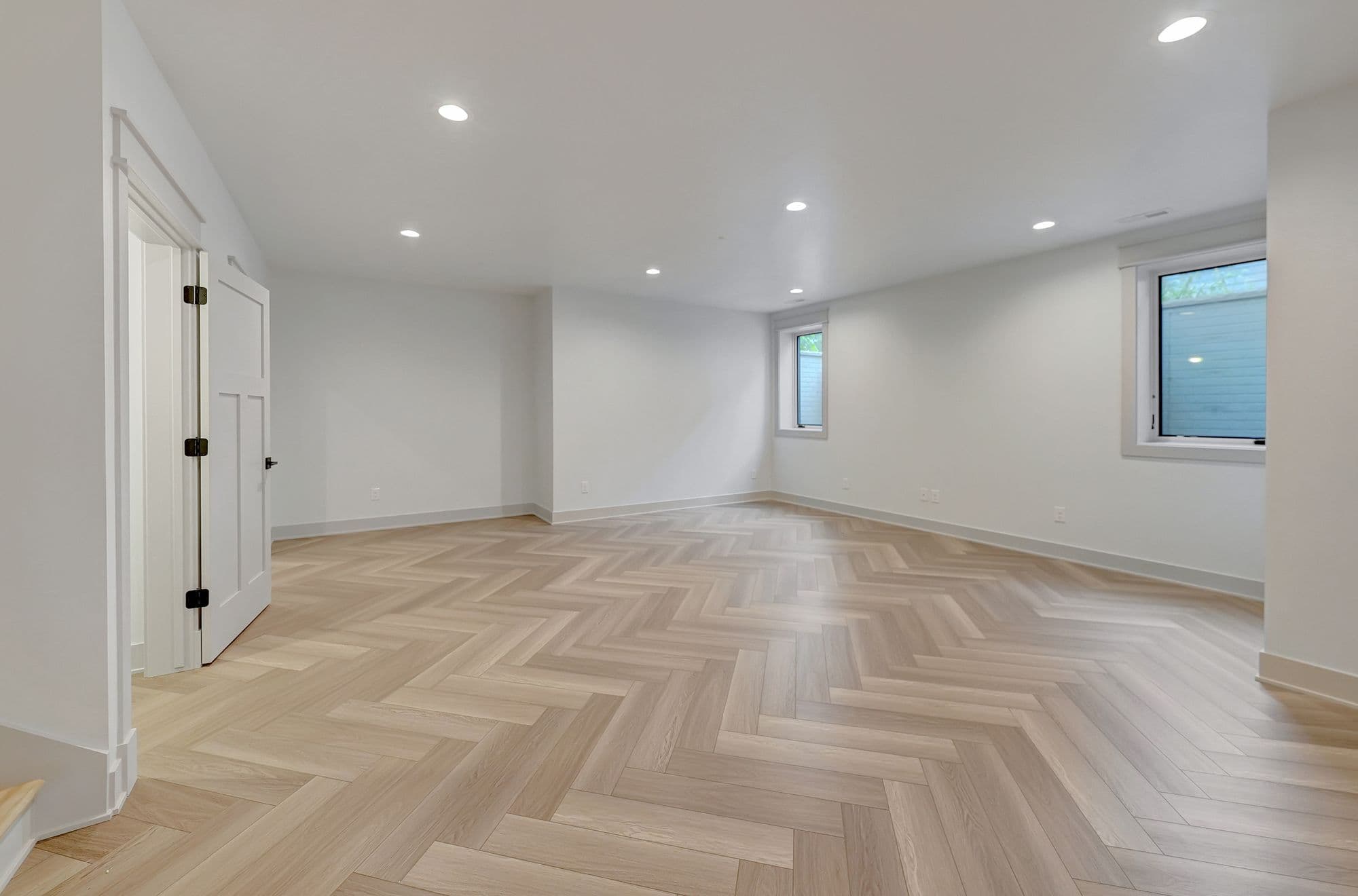 A brightly lit, spacious room features a herringbone-patterned wood floor and white walls. The room includes a white door, two windows, and recessed lighting, creating a clean and modern aesthetic.