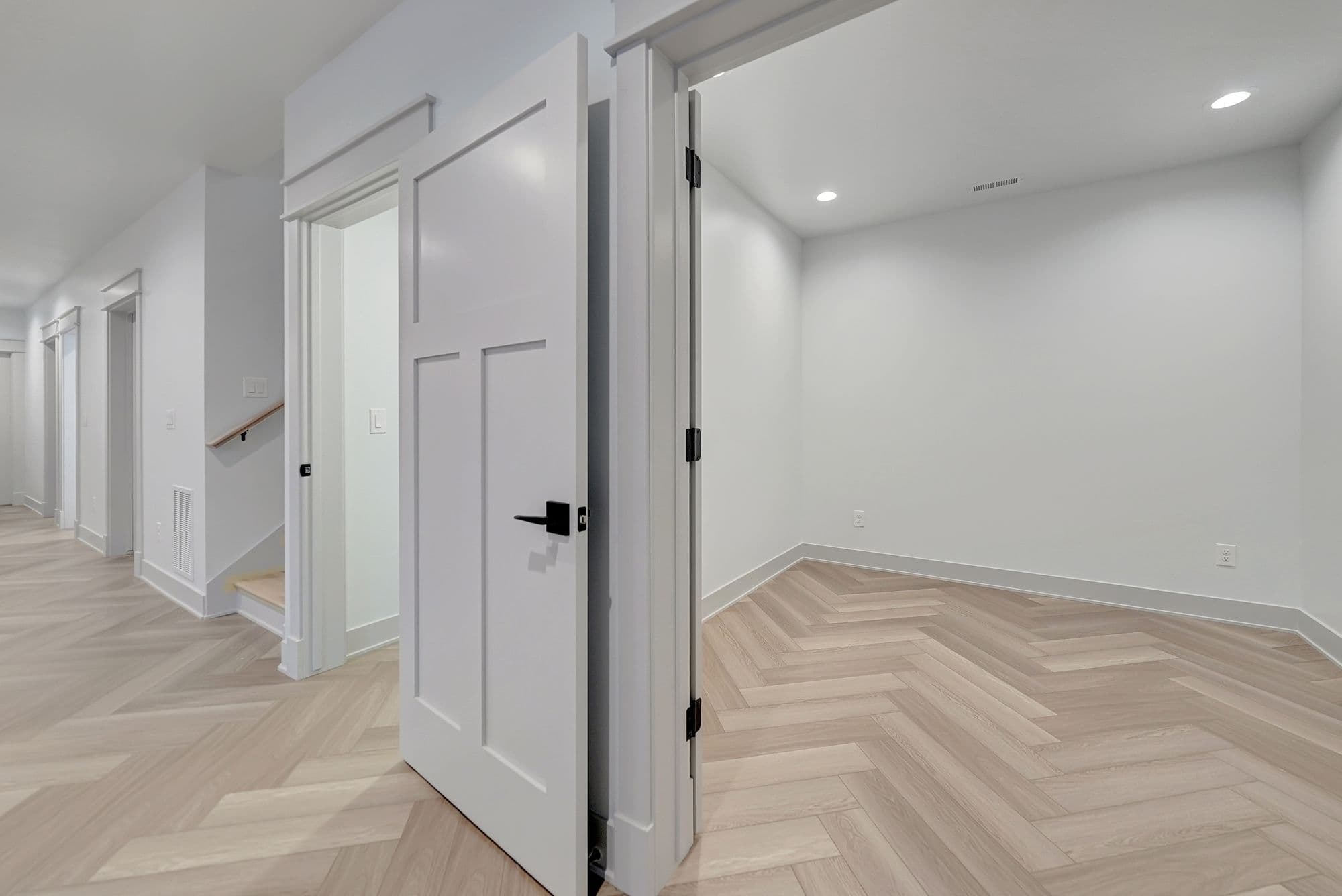 Bright entryway showcasing herringbone hardwood floors and white trim. An open doorway reveals a room with white walls, herringbone floors, and modern lighting.