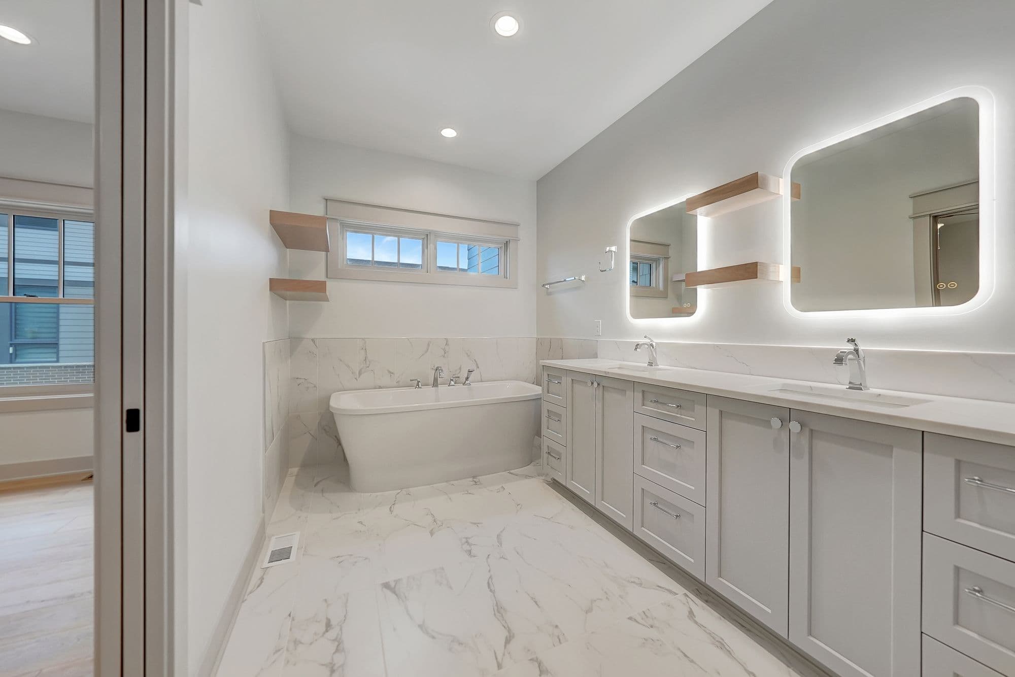 Modern bathroom featuring white marble flooring and wall tiling, a freestanding bathtub, and a gray double vanity with lighted mirrors and wood shelving. The room has a clean, bright aesthetic with a view to an exterior space.