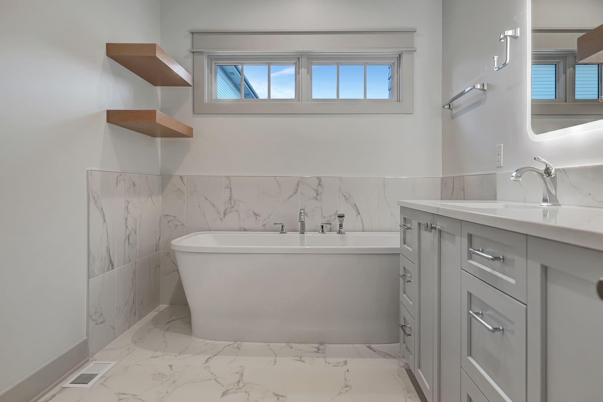 Bright, modern bathroom features a freestanding white soaking tub with chrome fixtures, marble-look tile walls and floor, floating wood shelves, and a gray vanity with white countertop and chrome hardware. The room is naturally lit by a window above the tub.