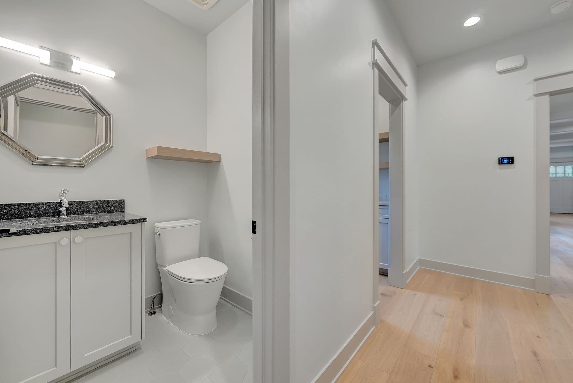 Powder room with white toilet, floating shelf, and cabinet vanity with black countertop. Hallway shows light wood flooring and door to an adjacent room.