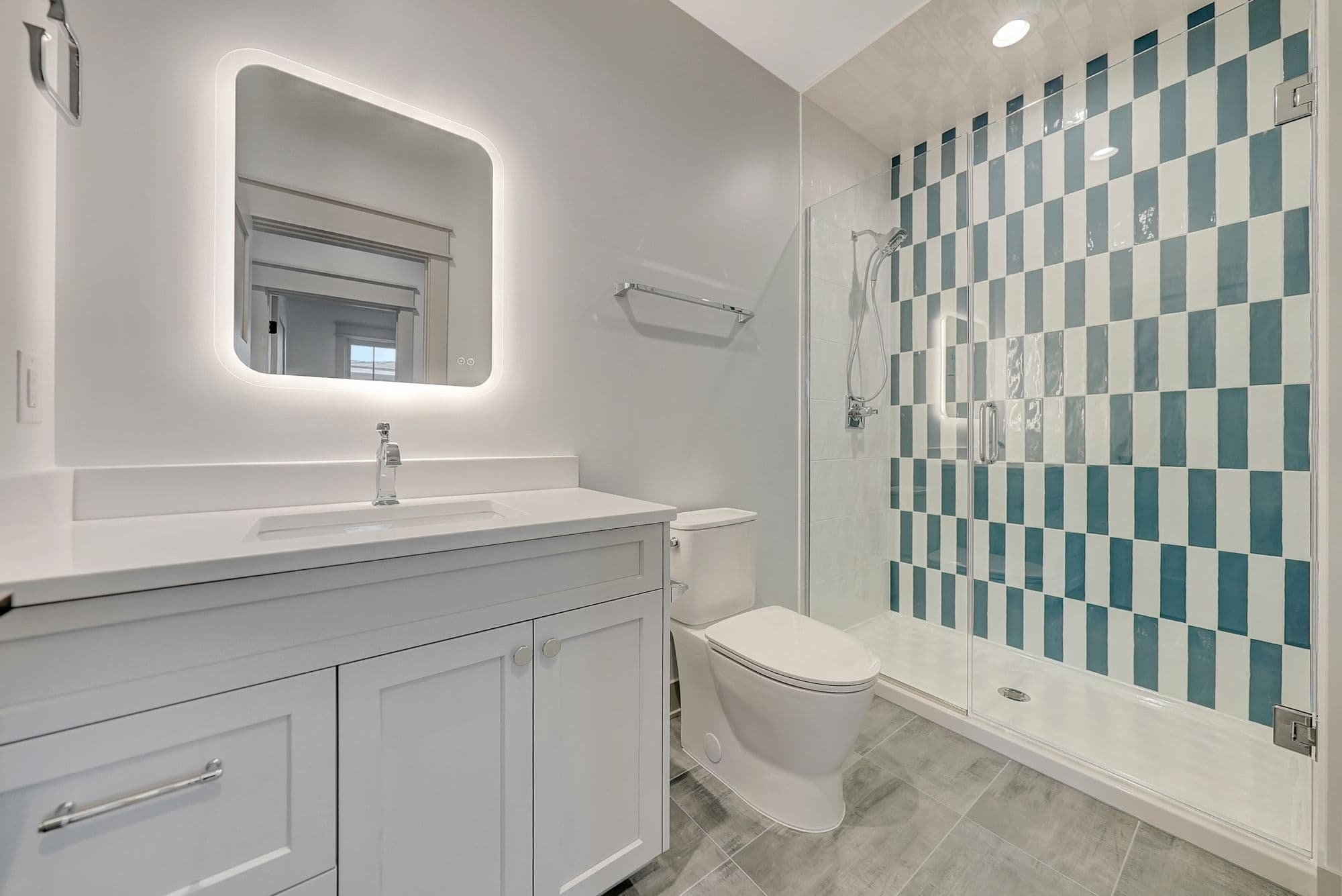 Bathroom features a modern white vanity with a lighted mirror, a white toilet, and a shower with teal and white vertical tiles behind a glass door. The floor is covered in gray tiles.