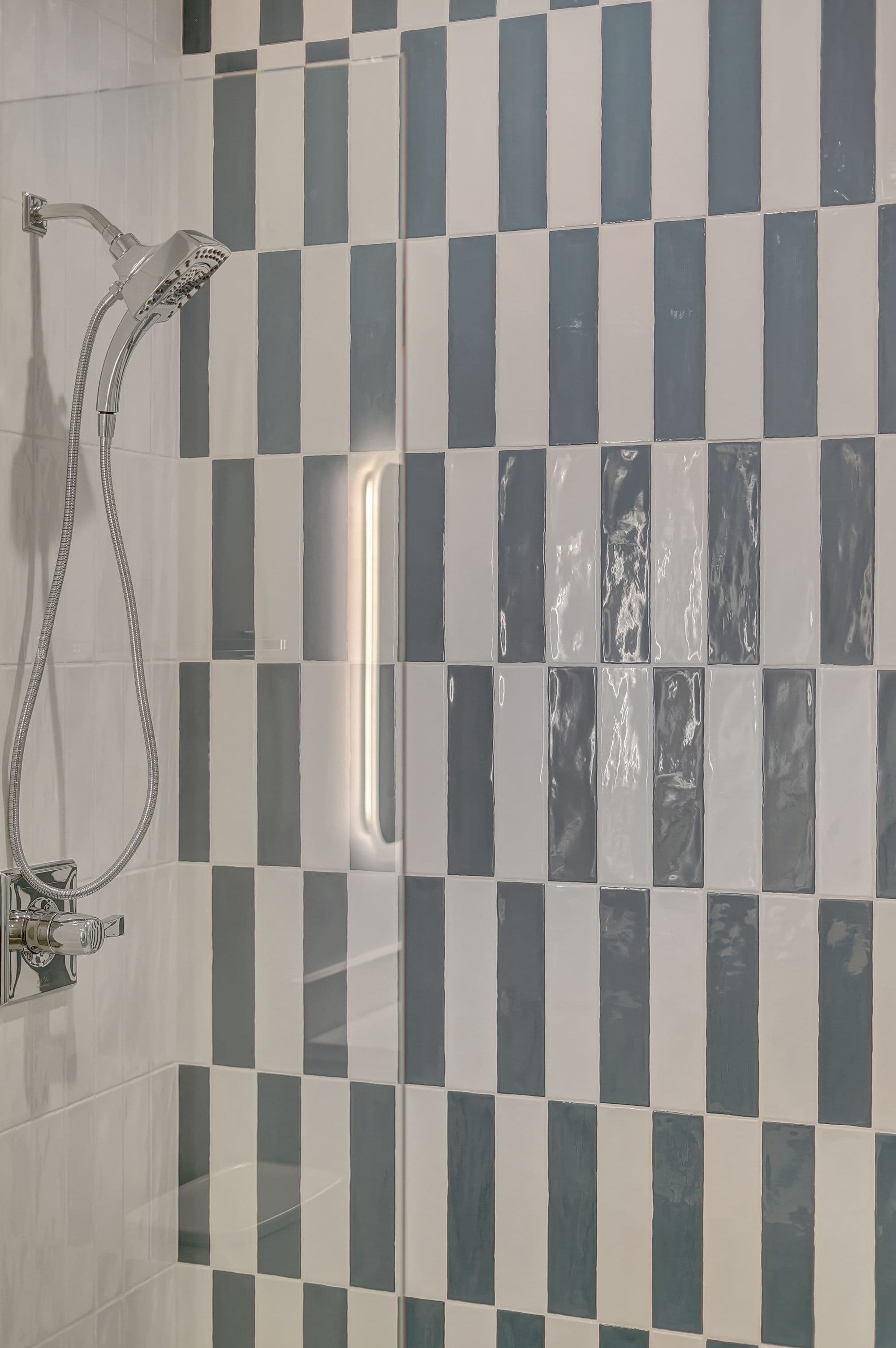 Close-up of a modern bathroom shower area featuring a vertically striped tile pattern in white and gray. The shower head and handle are chrome, and there is a clear glass enclosure.