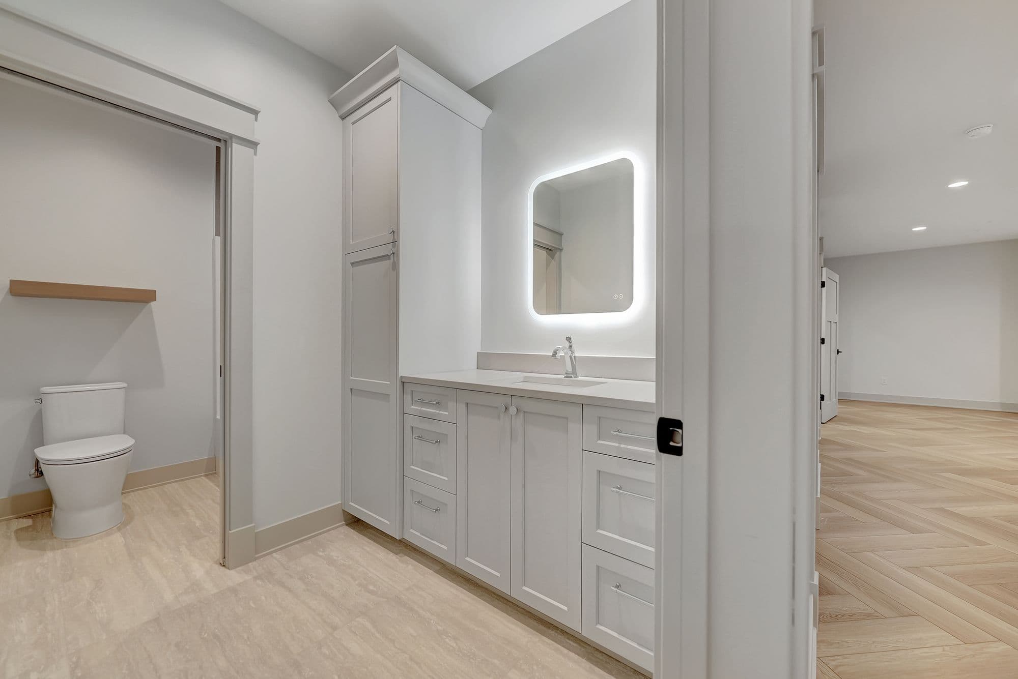 Modern bathroom featuring a lighted vanity mirror and white cabinetry with drawers and cabinets. A toilet is visible through an open doorway, and herringbone wood floors extend into an adjacent room.
