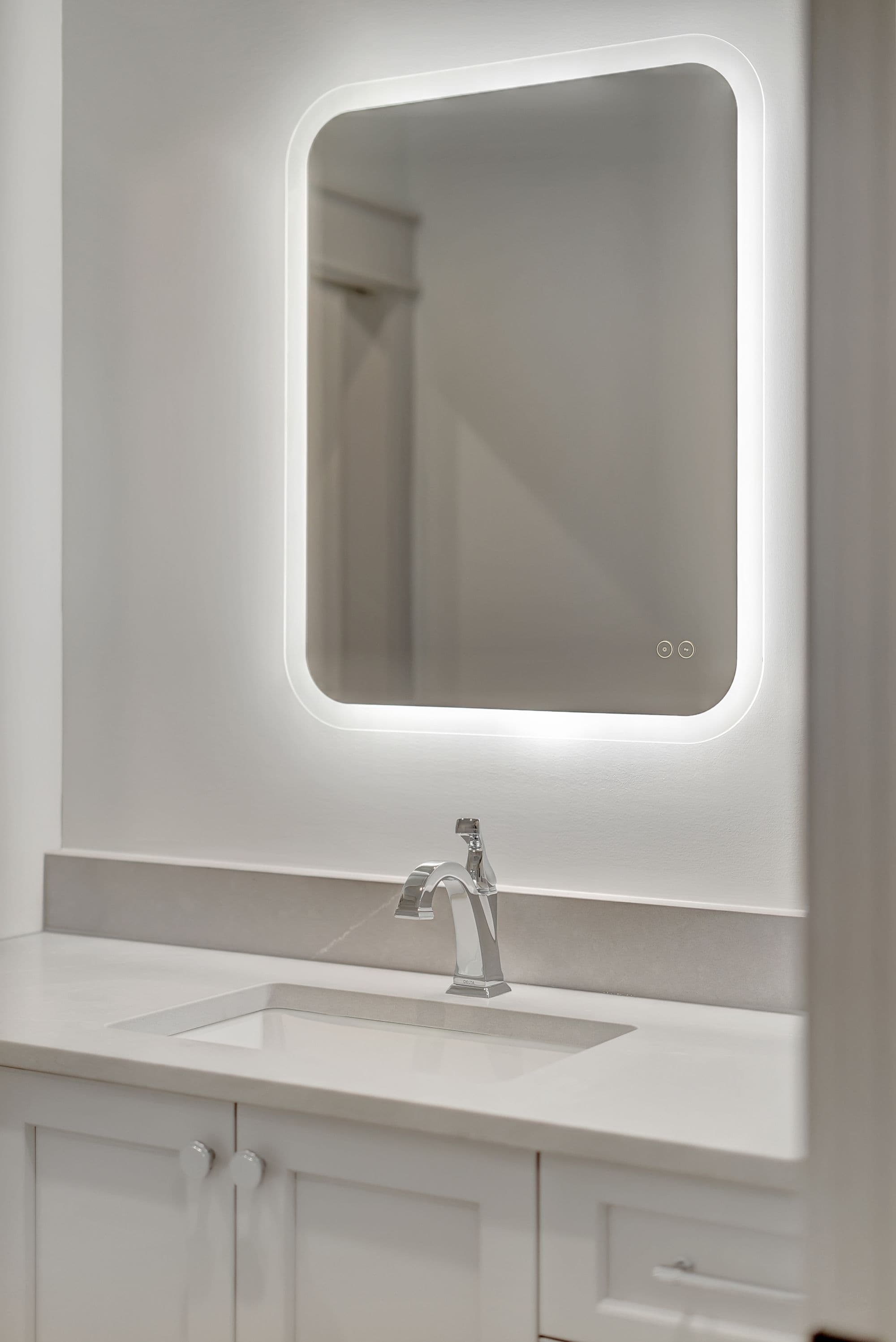 Bathroom vanity featuring a white countertop, a rectangular sink, and a chrome faucet is seen. An LED-backlit mirror with rounded corners hangs above the vanity.