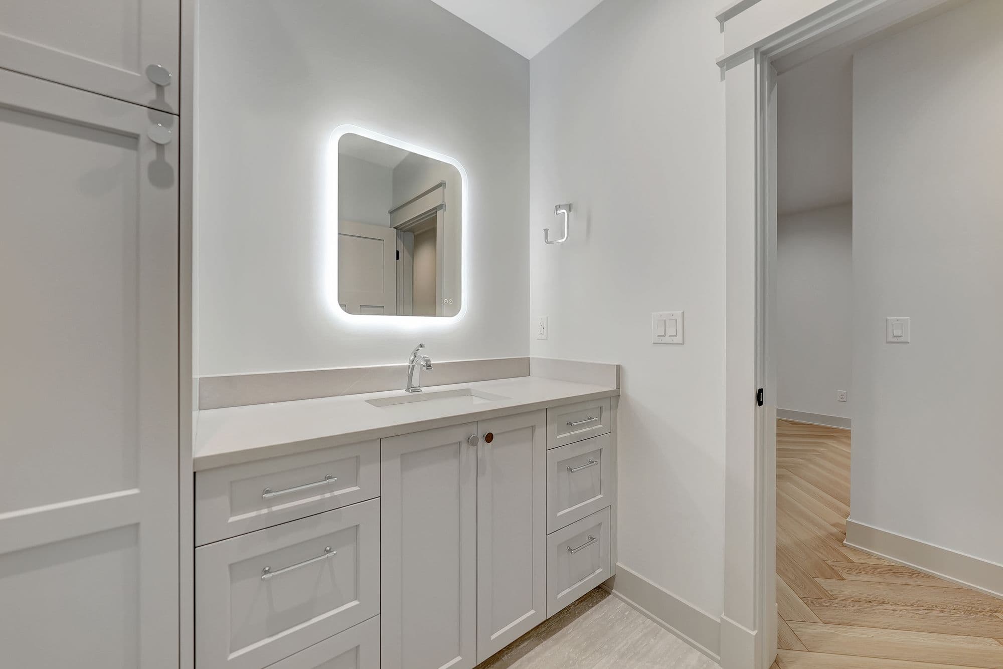 Modern bathroom features a white vanity with light countertops, a rounded rectangular mirror with built-in lighting, and light gray walls. The room opens to another space with hardwood floors laid in a herringbone pattern.