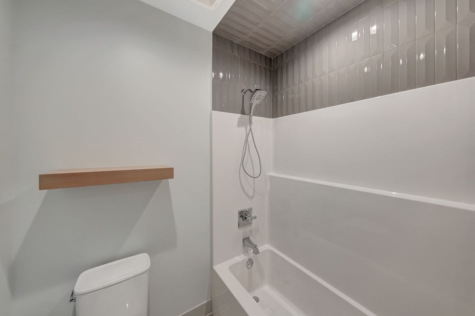 Bathroom features a modern design with gray vertically-stacked tiles above a white tub/shower combination. A light wood floating shelf sits above a white toilet against a light gray wall.
