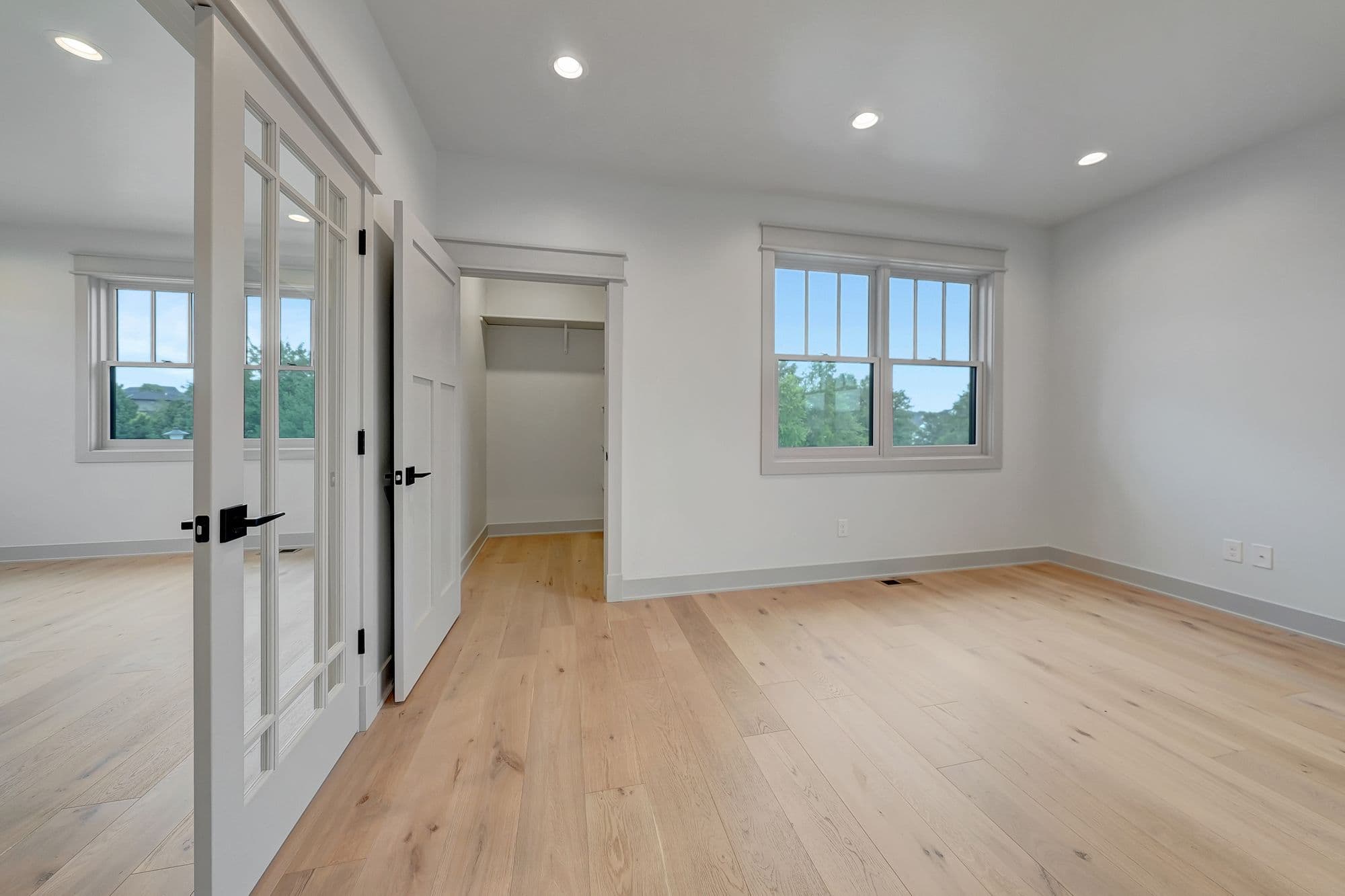A bedroom features light hardwood flooring, white walls, and neutral gray trim. A pair of glass-paneled doors open into the room, with a closet and window visible in the background.