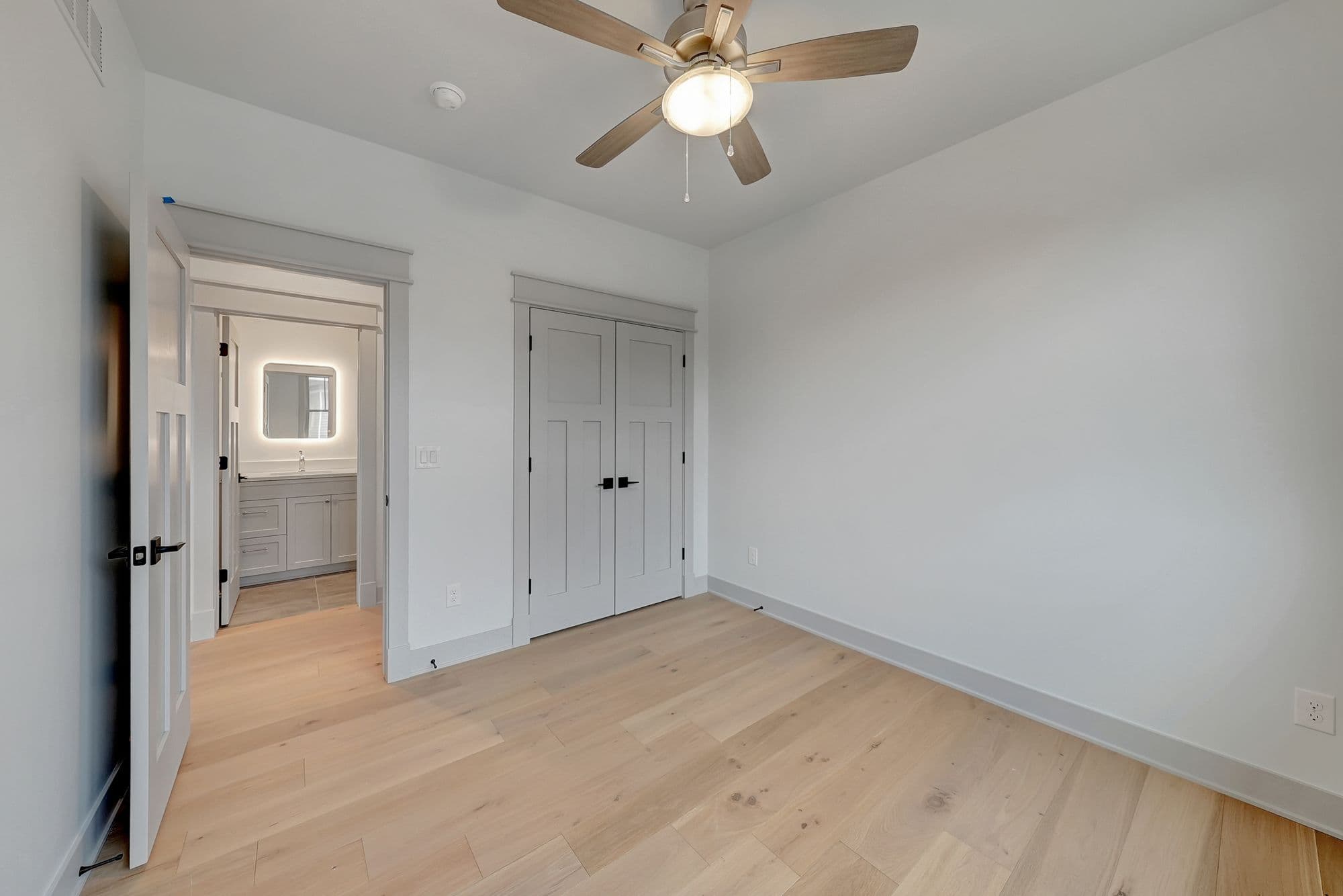 Bedroom featuring light hardwood floors, white walls, and a ceiling fan with a light fixture. A doorway leads to a bathroom with a vanity and mirror.