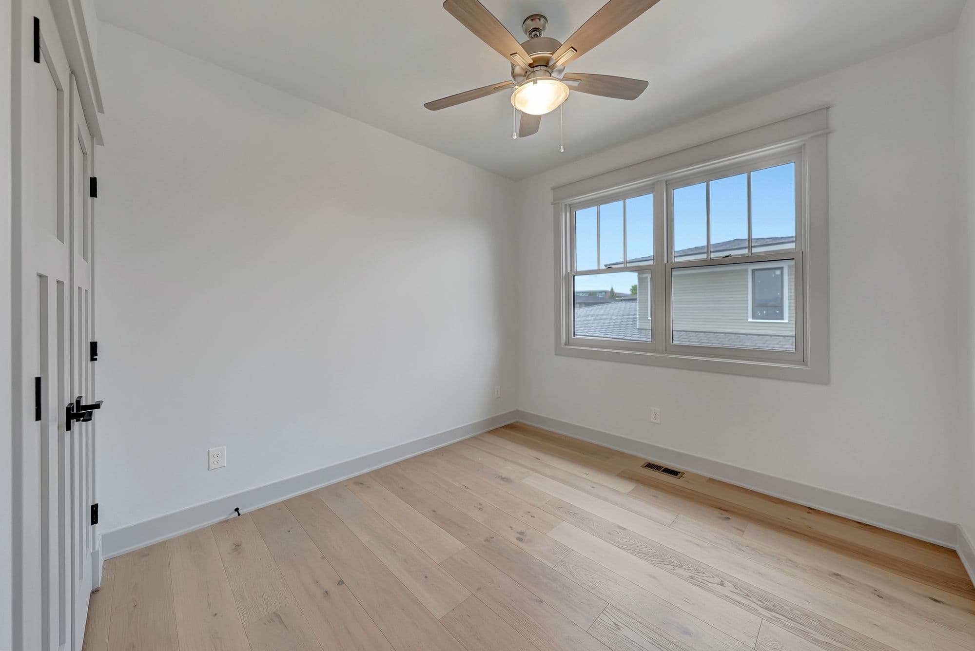 Bright bedroom with light hardwood floors, white walls, and a ceiling fan. The room has a window with a view of nearby houses.