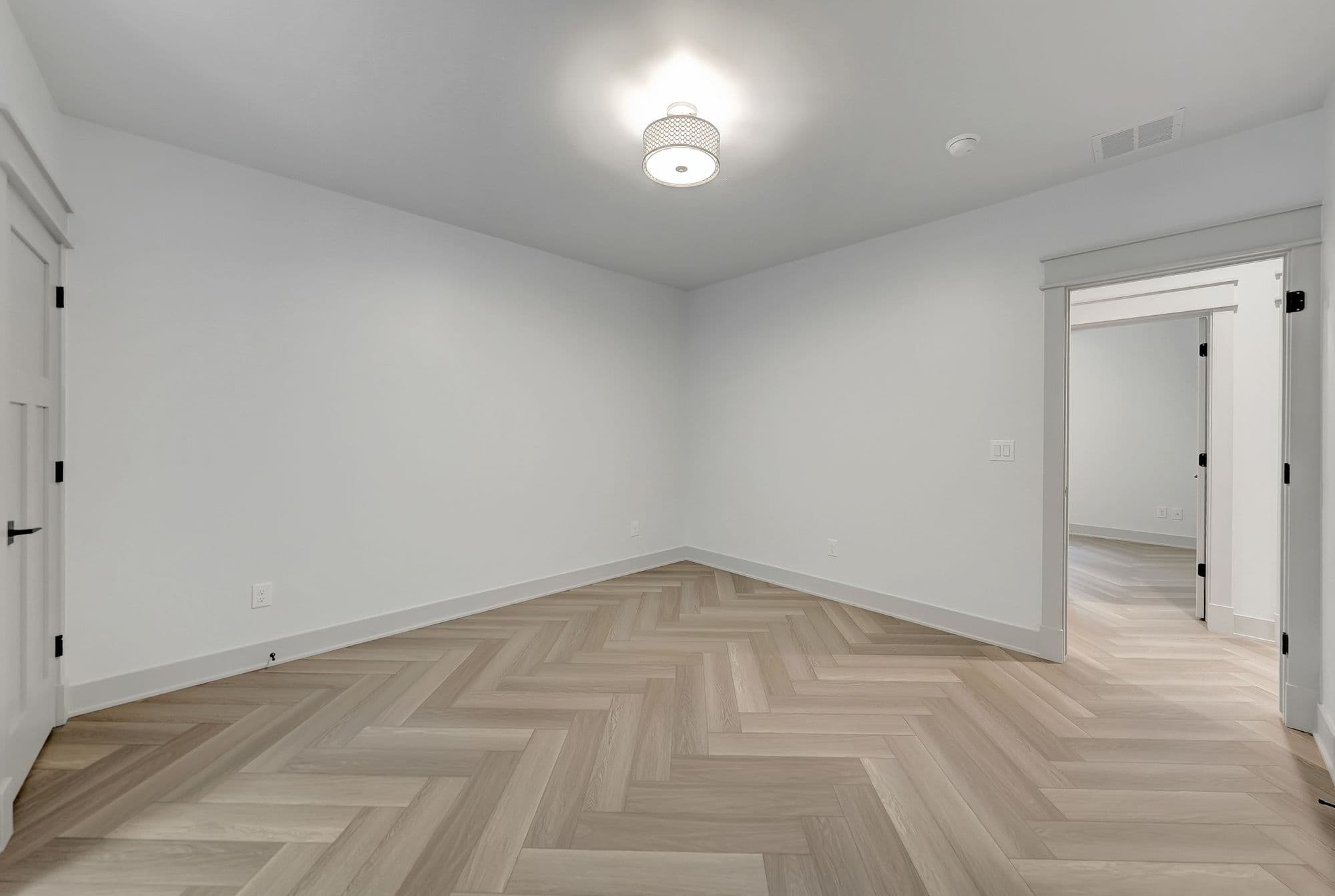 Bright, modern room with herringbone pattern hardwood floors and white walls and trim. A doorway leads to another room with matching floors.