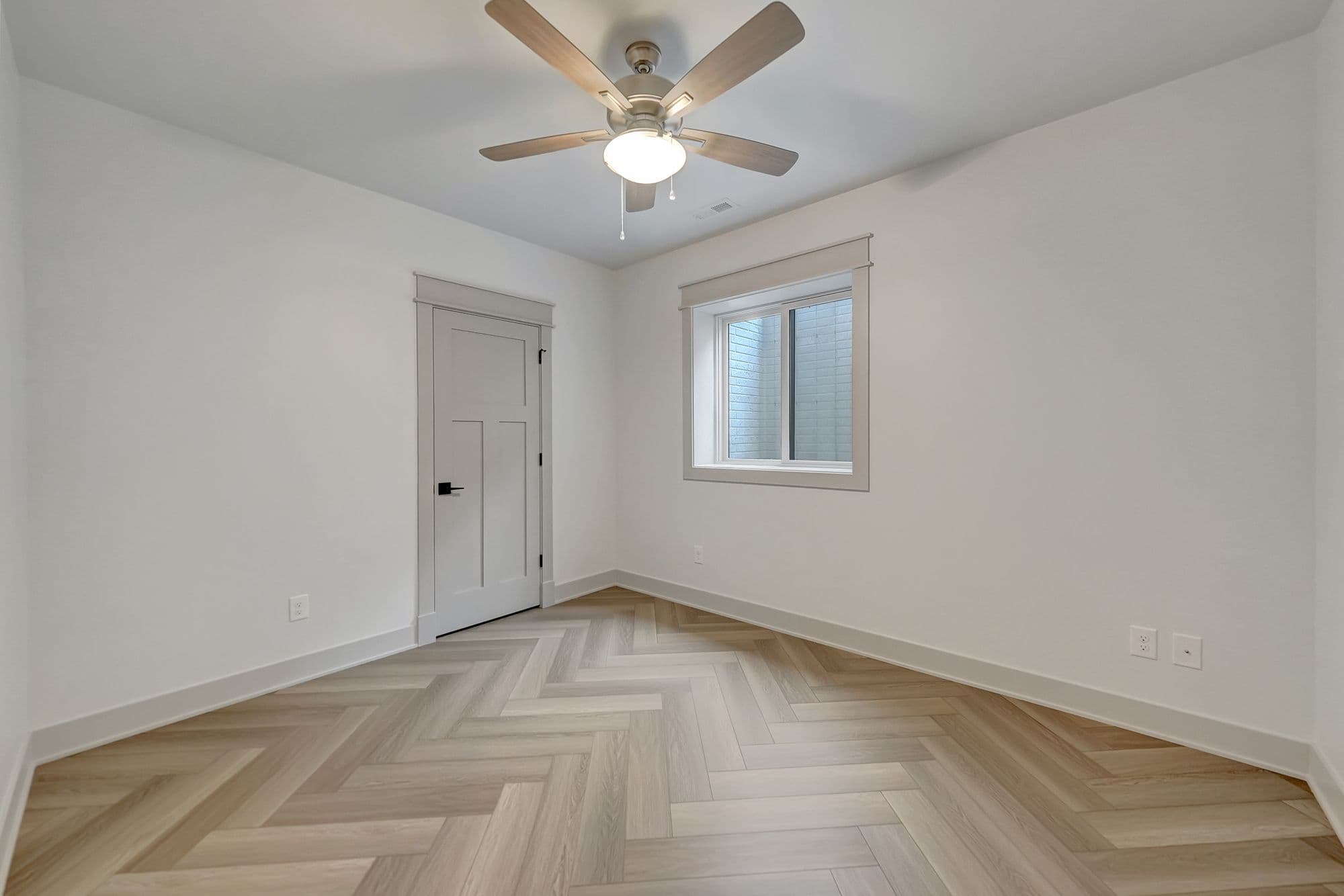 A bedroom is shown with a light wood herringbone floor, light gray door and trim, and white walls. A modern ceiling fan with five blades and a light fixture is centered in the room.