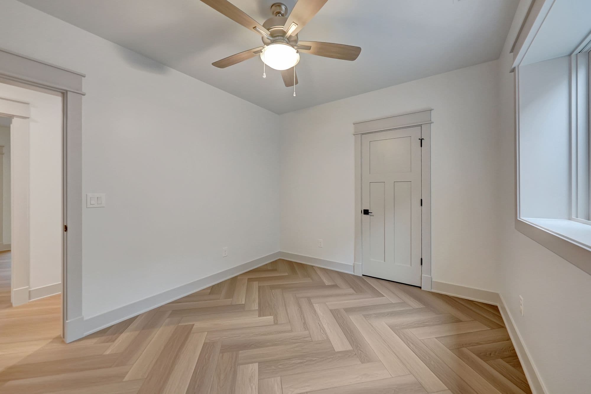 Bright bedroom with herringbone wood floors, neutral walls, and a ceiling fan with a light. A doorway leads out of the room, and another interior door is visible on the right wall.