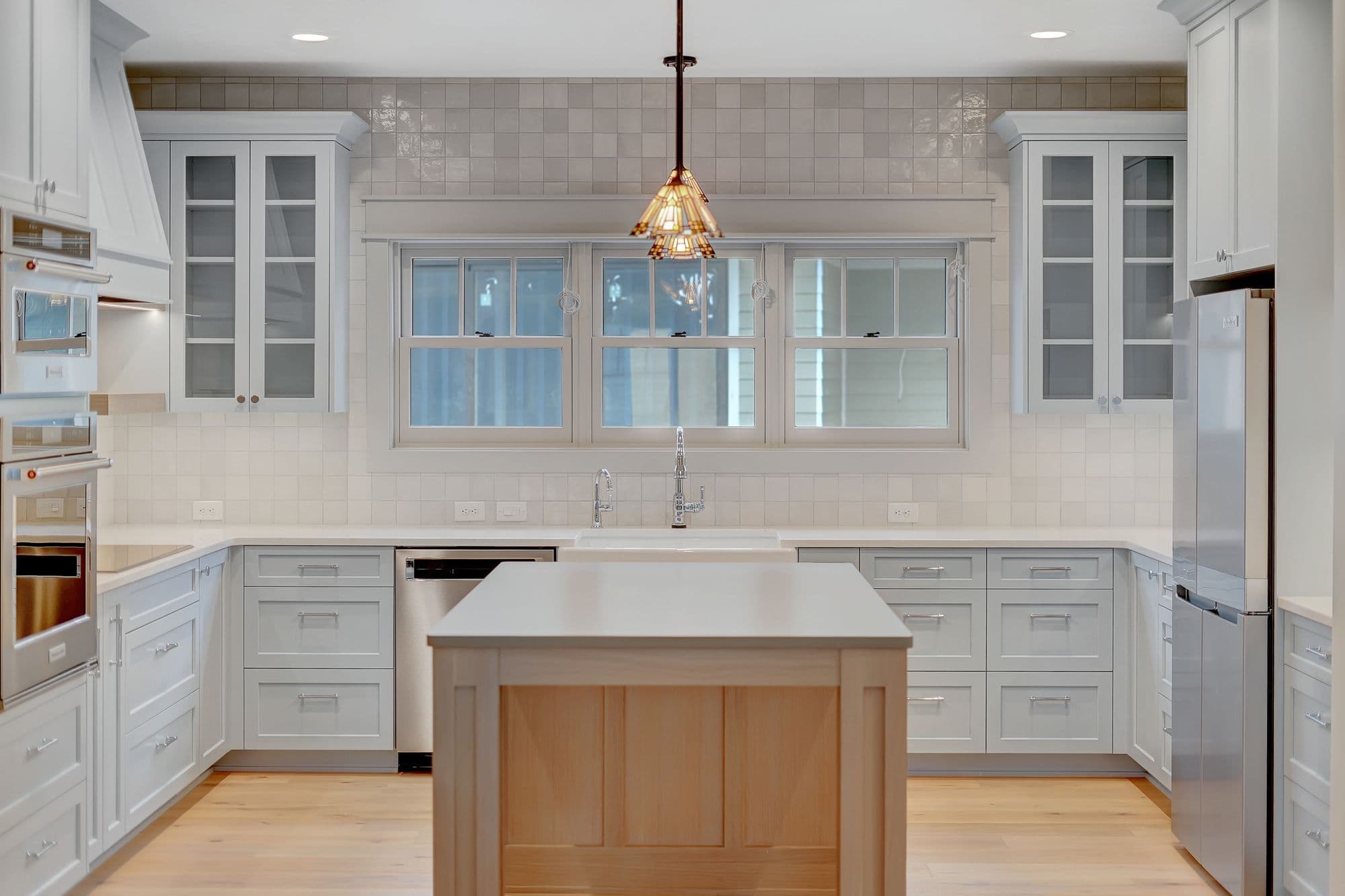 Well-lit kitchen features white cabinets, stainless steel appliances, and a kitchen island with a light wood base. A modern light fixture hangs above the island, and a window above the sink provides natural light.