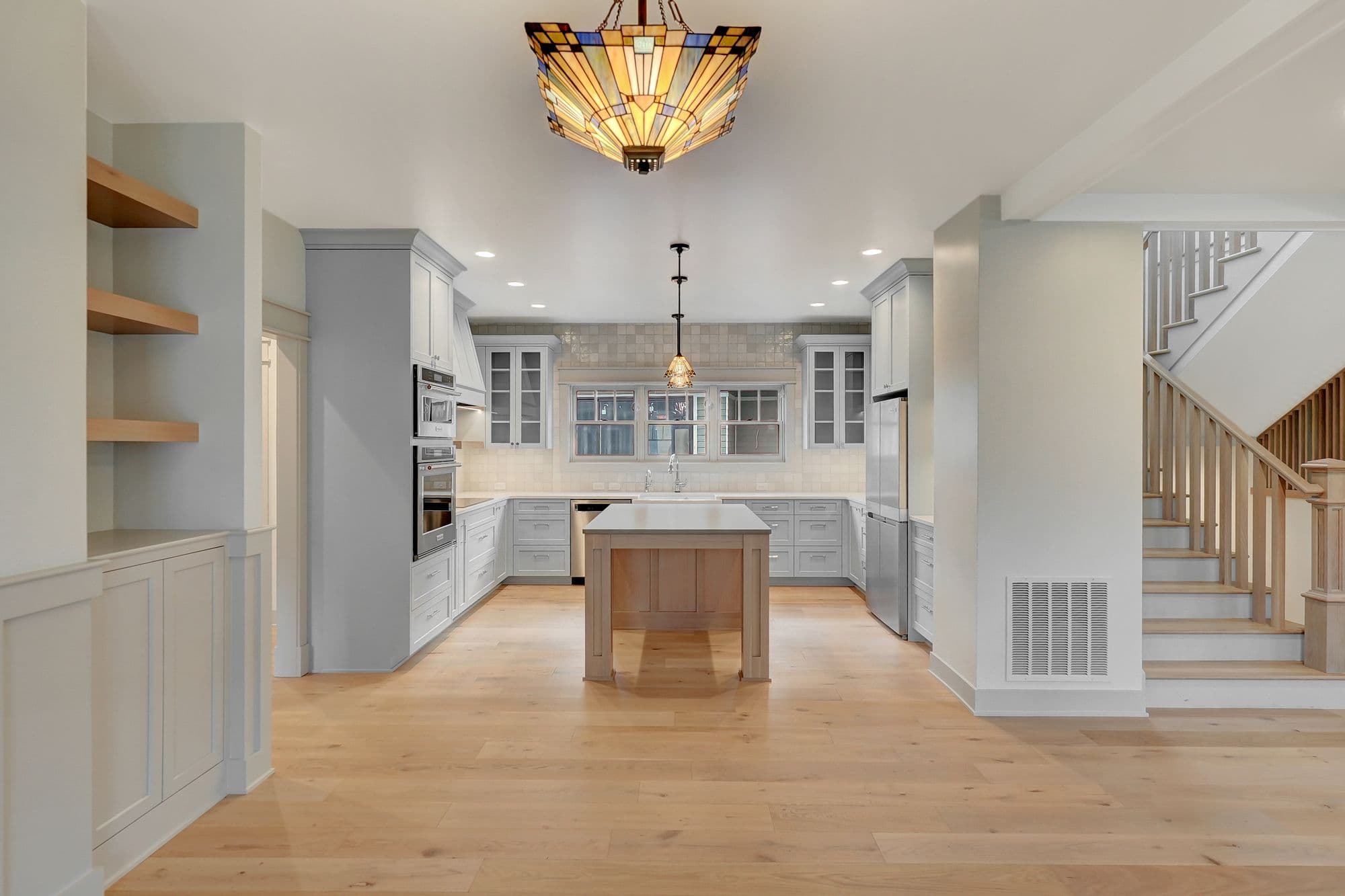 Bright, open kitchen features gray cabinetry, wood floors, and a kitchen island. A stained glass light fixture adds a pop of color above the island and natural light filters in through the windows above the kitchen sink.