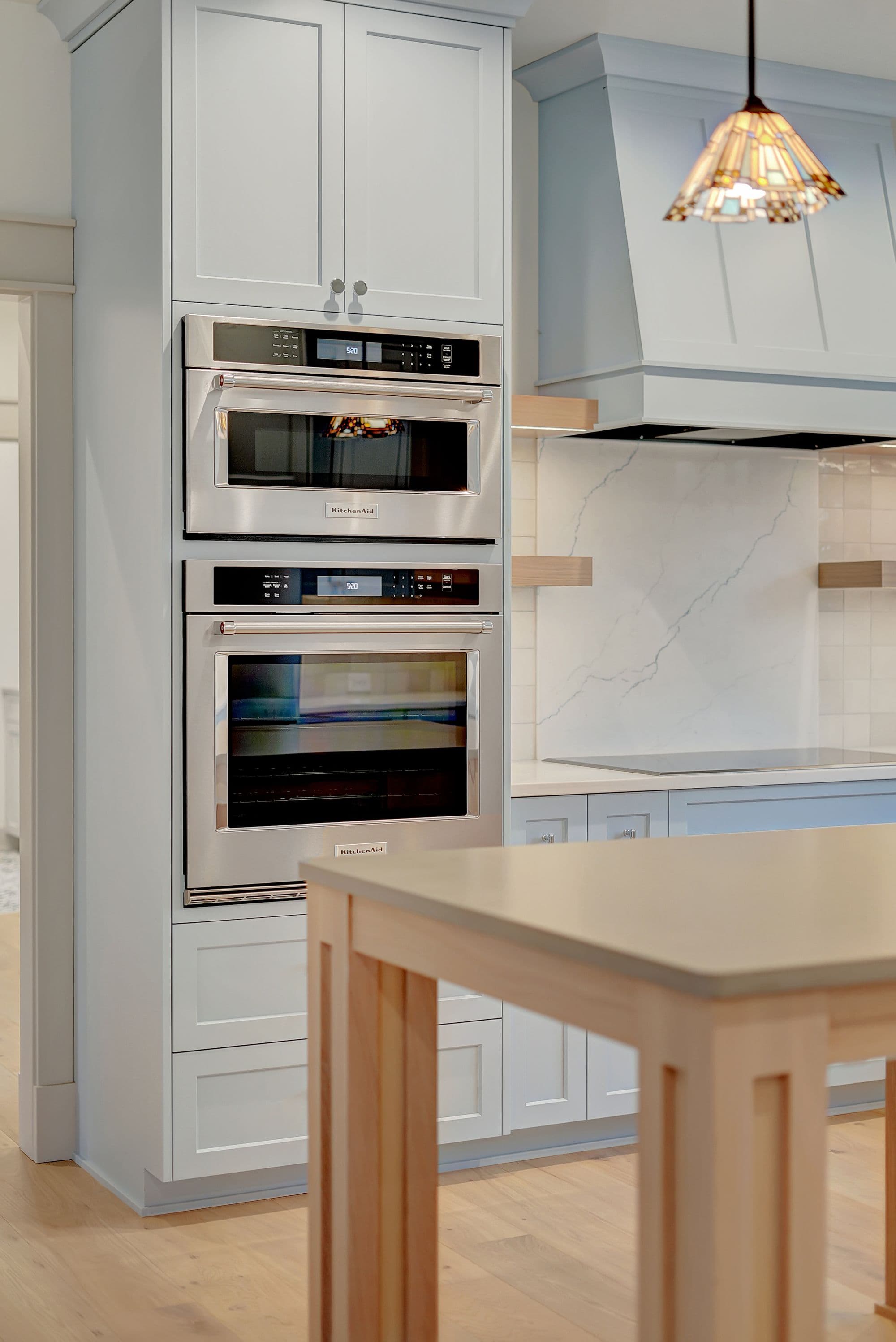 Modern kitchen features built-in KitchenAid stainless steel microwave and oven appliances with light blue cabinetry. A light wood island sits in front of the appliances, with a decorative range hood and white tile backsplash in the background.